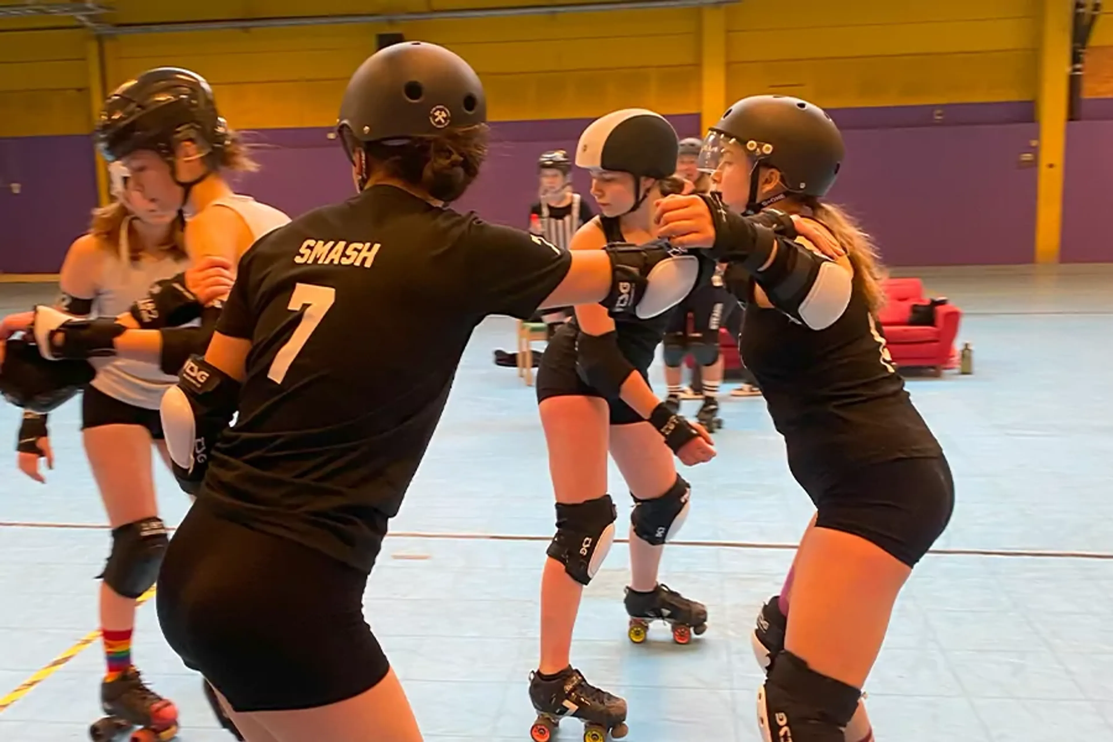 Fyra rullskridskoåkare i skyddsutrustning tränar tillsammans inomhus på en bana. Två av dem bär svart matchande kläder med nummer på ryggen.