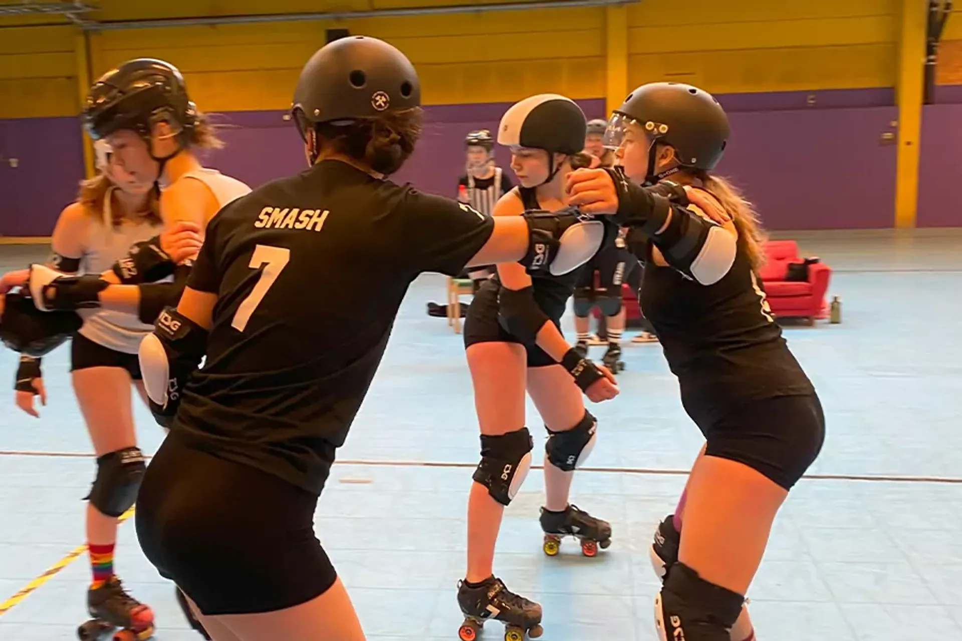 Fyra rullskridskoåkare i skyddsutrustning tränar tillsammans inomhus på en bana. Två av dem bär svart matchande kläder med nummer på ryggen.