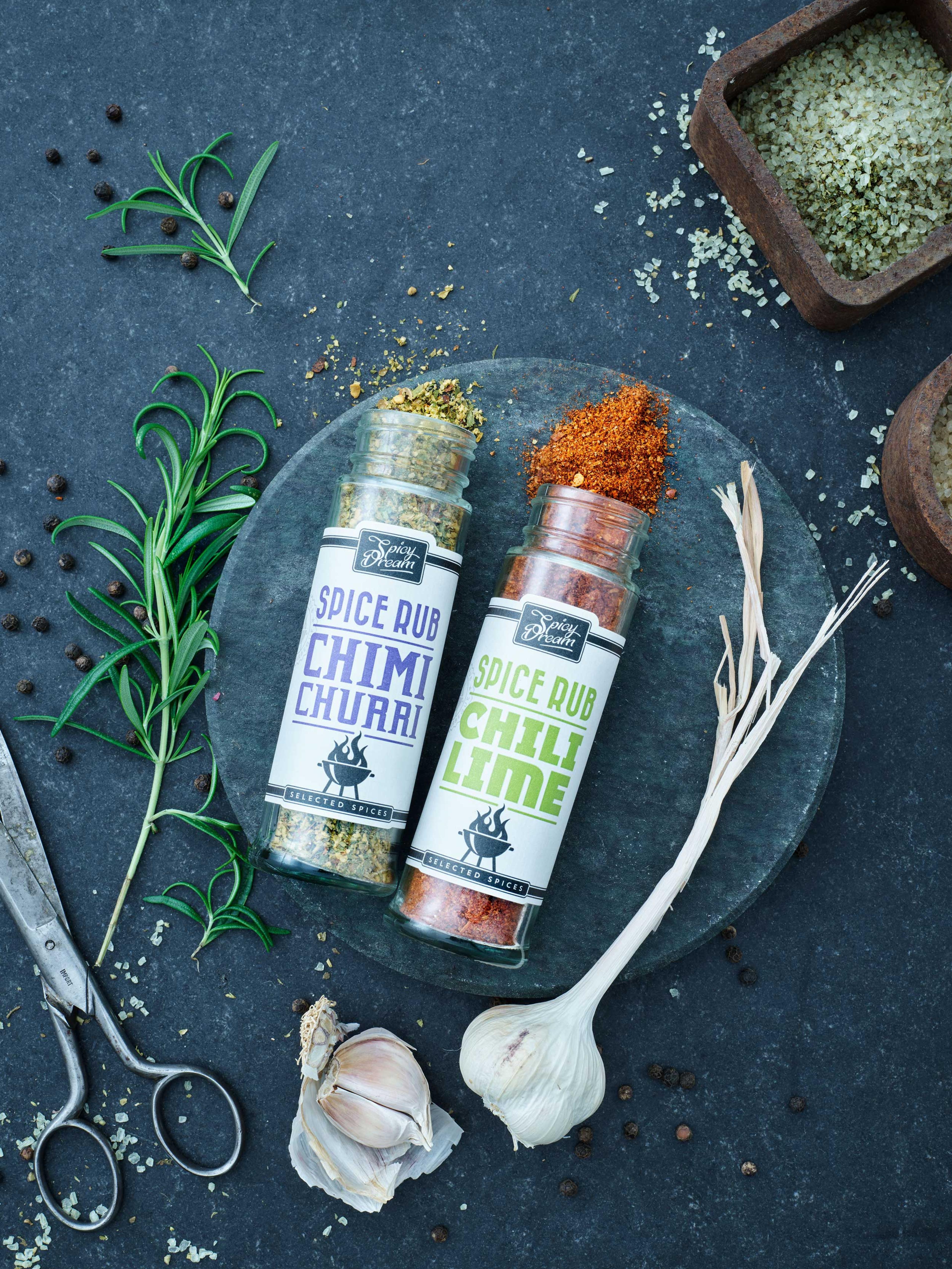 Två kryddburkar med rub-smaker, en med chimi churri och en med chili lime, placerade på ett mörkt underlag tillsammans med färsk vitlök, rosmarin och en kökssax. Små högar av kryddblandningar och