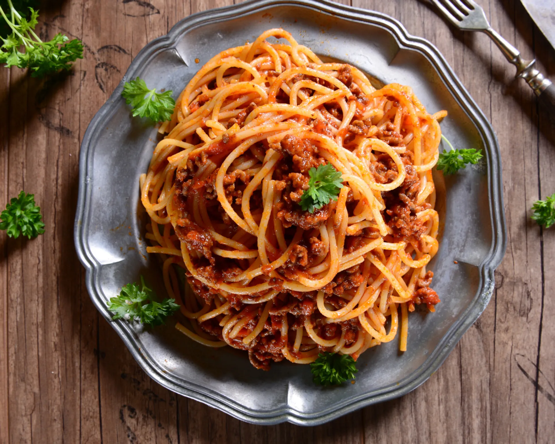 En tallrik med spaghetti och köttfärssås, garnerad med färsk persilja. Serverad på en äldre, metallgrå tallrik på ett träbord.