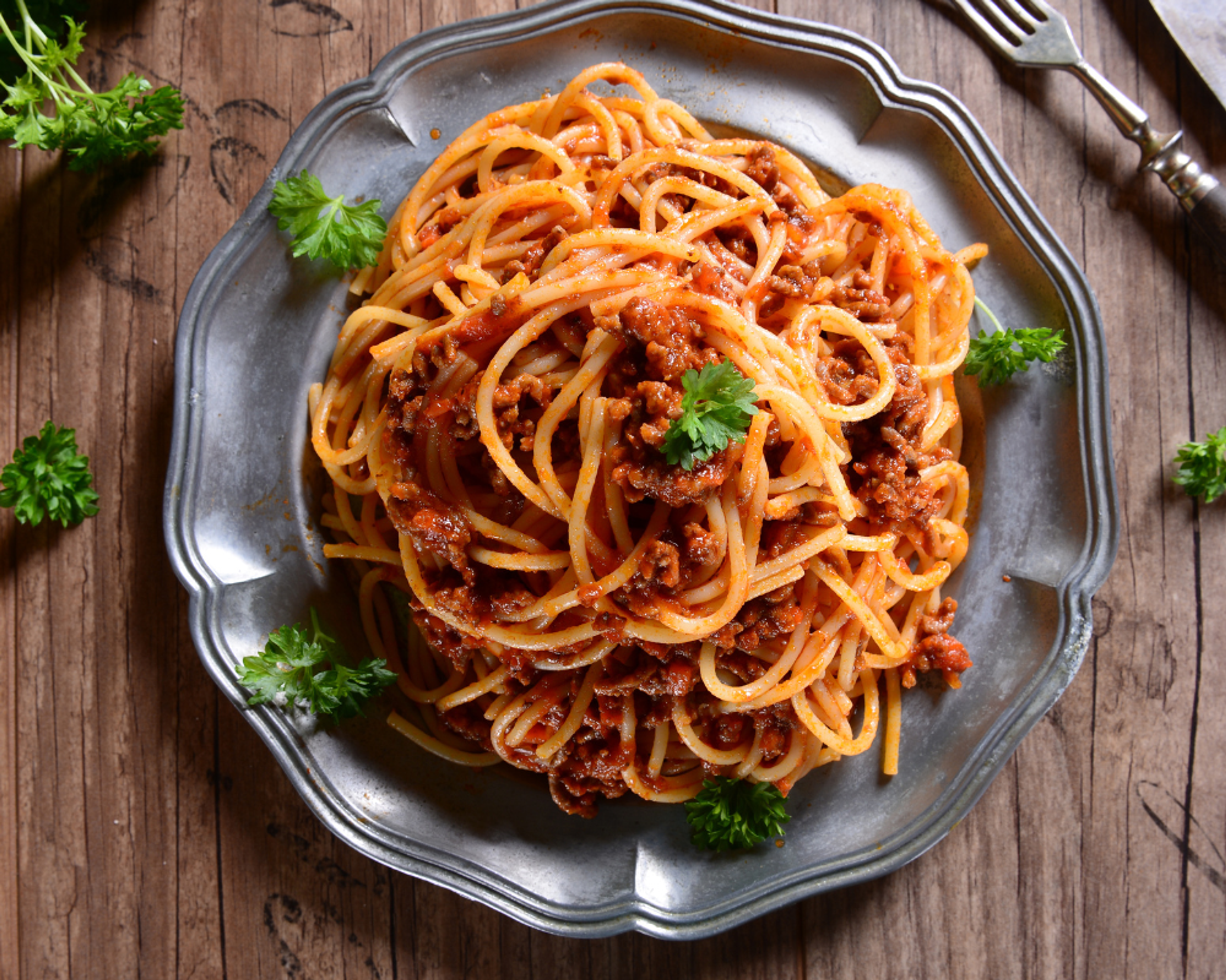 En tallrik med spaghetti och köttfärssås, garnerad med färsk persilja. Serverad på en äldre, metallgrå tallrik på ett träbord.