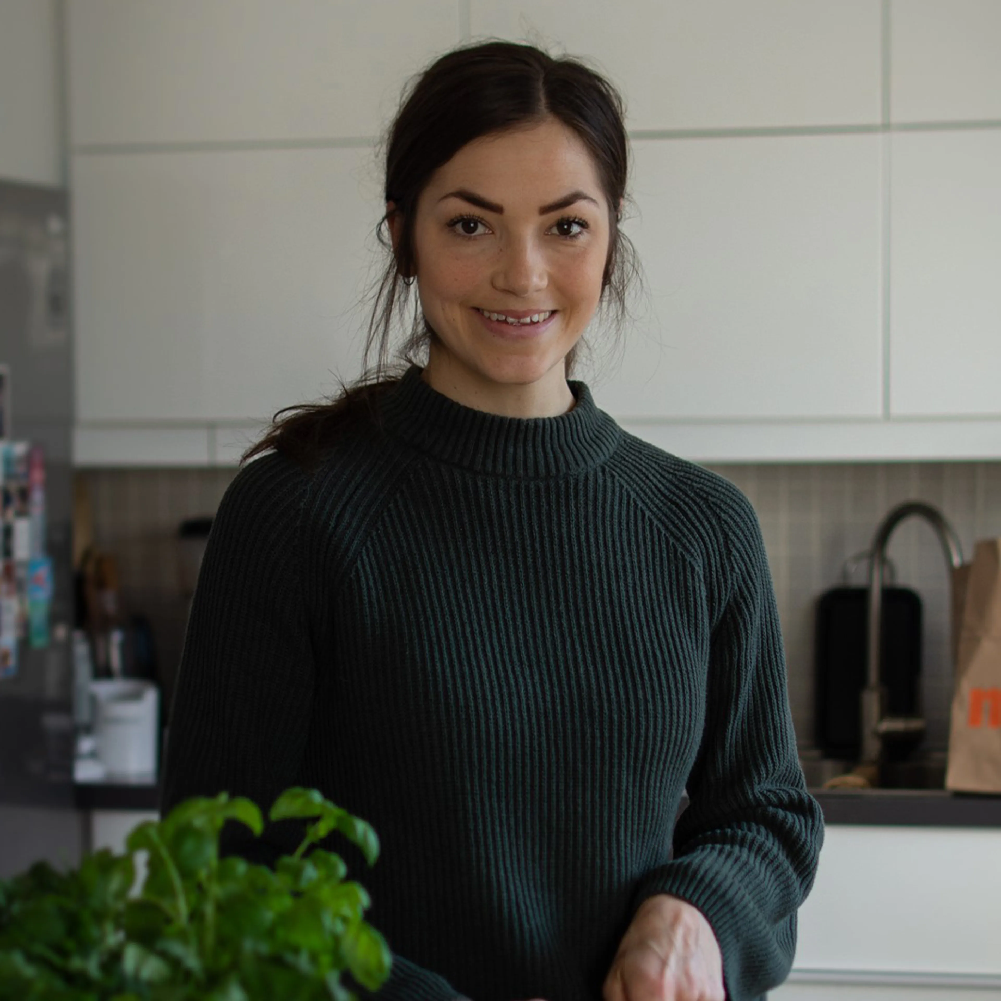 En kvinna med mörkt hår i en grön ribbstickad tröja står i ett ljust kök med vit inredning. Framför henne syns en grön växt, och hon ler mot kameran.