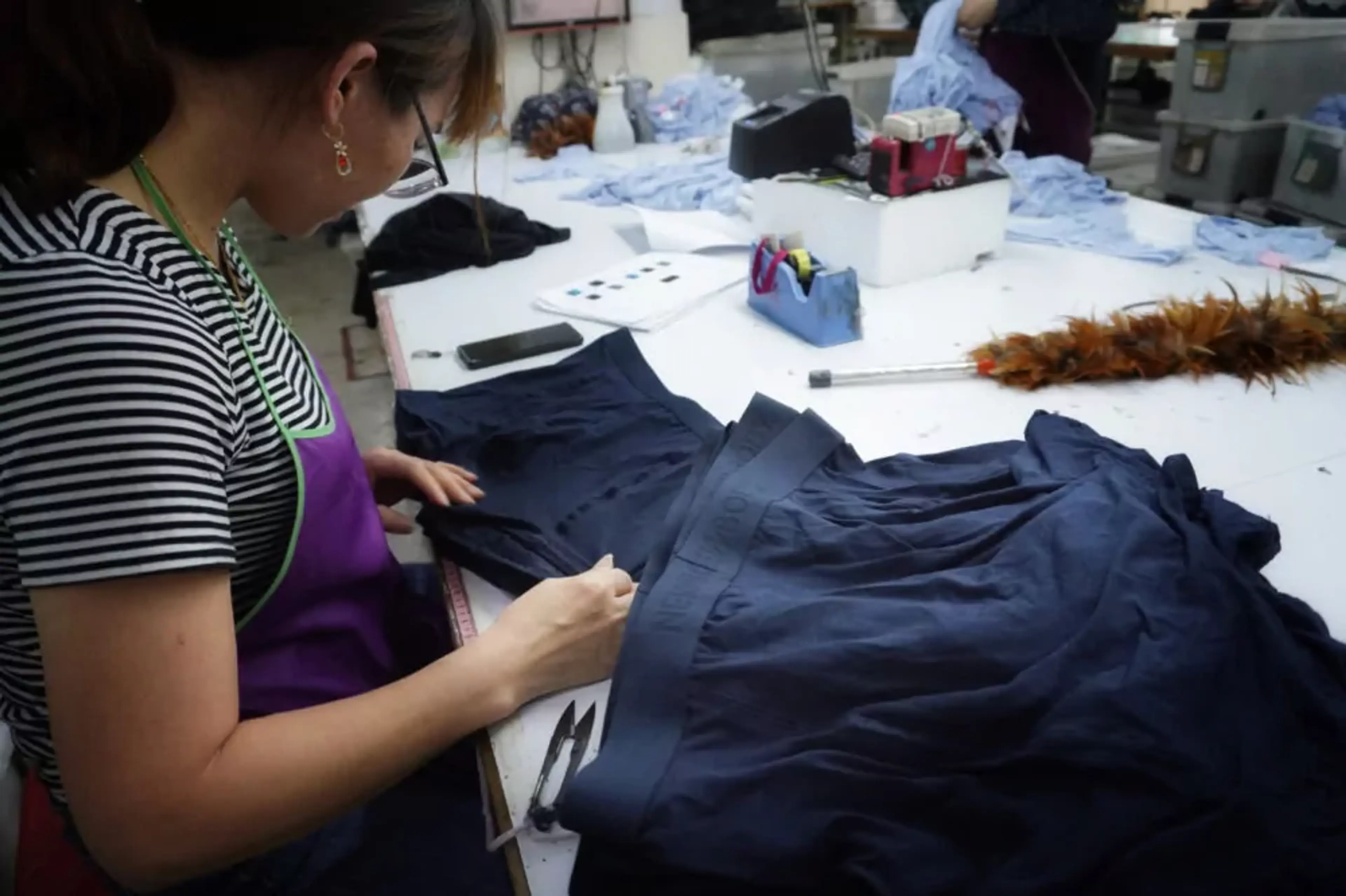 En person granskar och arbetar med mörkblå textilmaterial på ett bord i en produktionsmiljö. Omkring ligger olika arbetsverktyg och annat tyg.