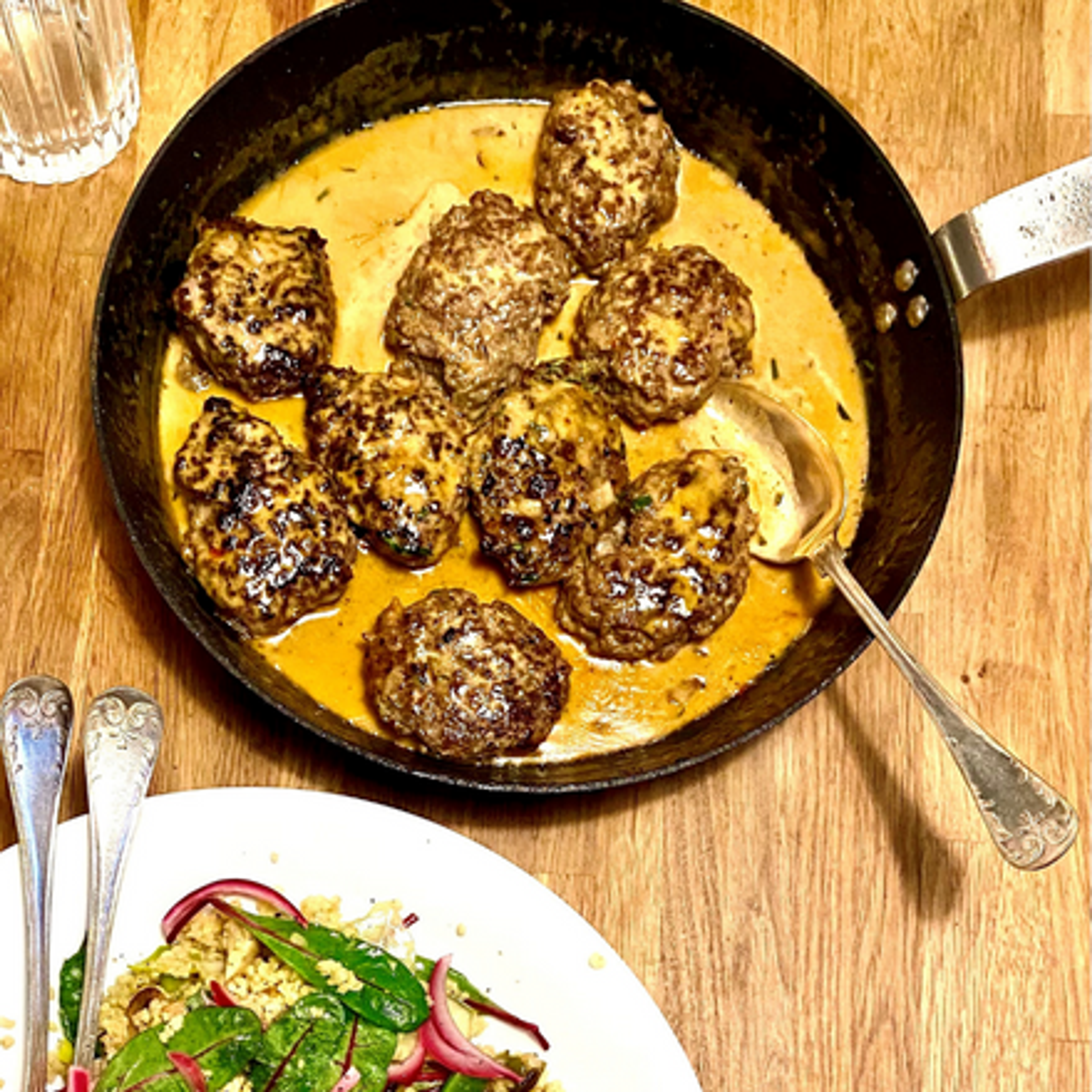 Färsbiffar som steks i en panna med krämig sås, serverade på ett träbord. En tallrik med sallad syns i förgrunden.