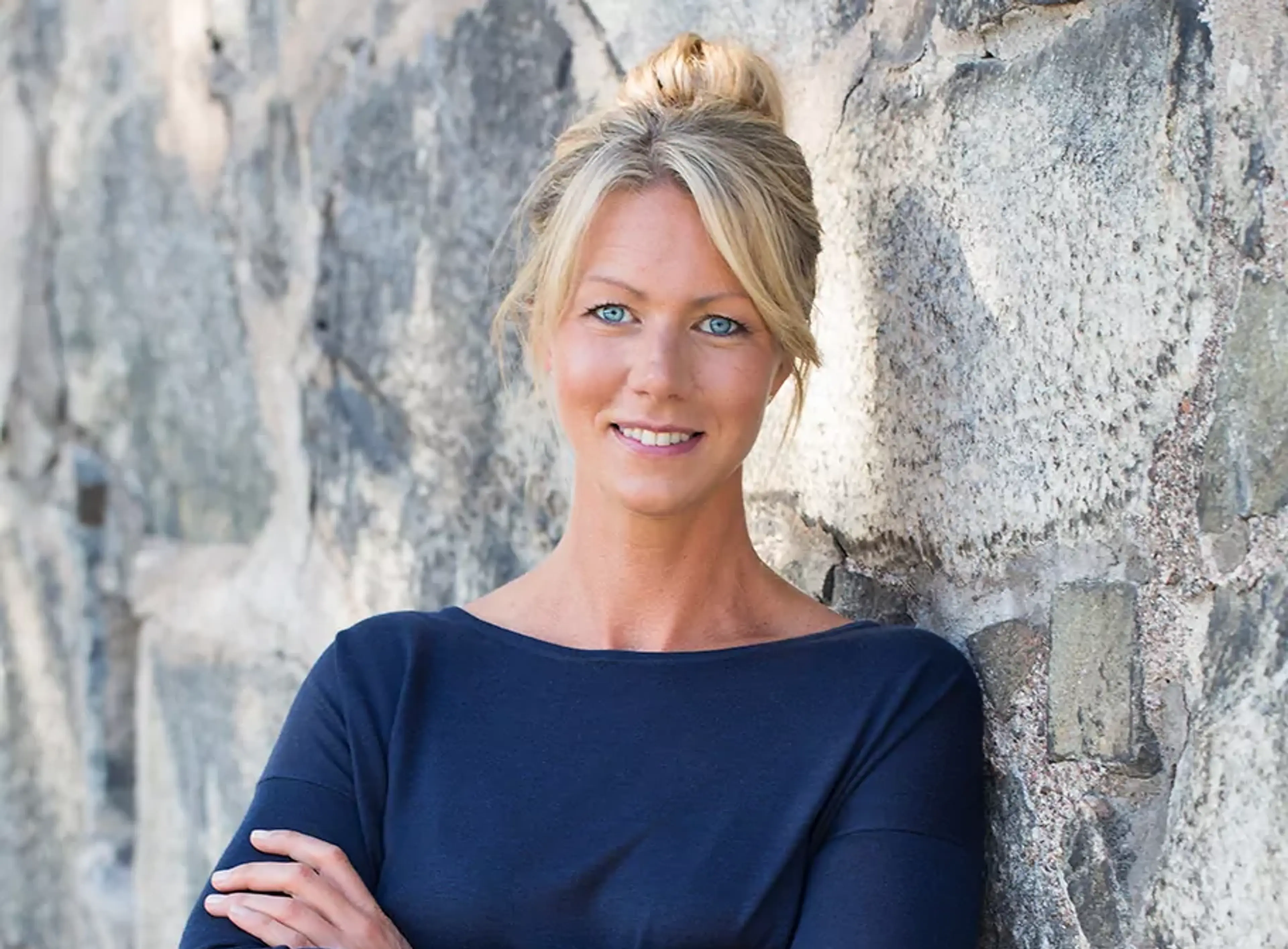 Kvinna med ljusblont hår uppsatt i knut, klädd i en mörkblå tröja, står lutad mot en stenmur med armarna i kors.