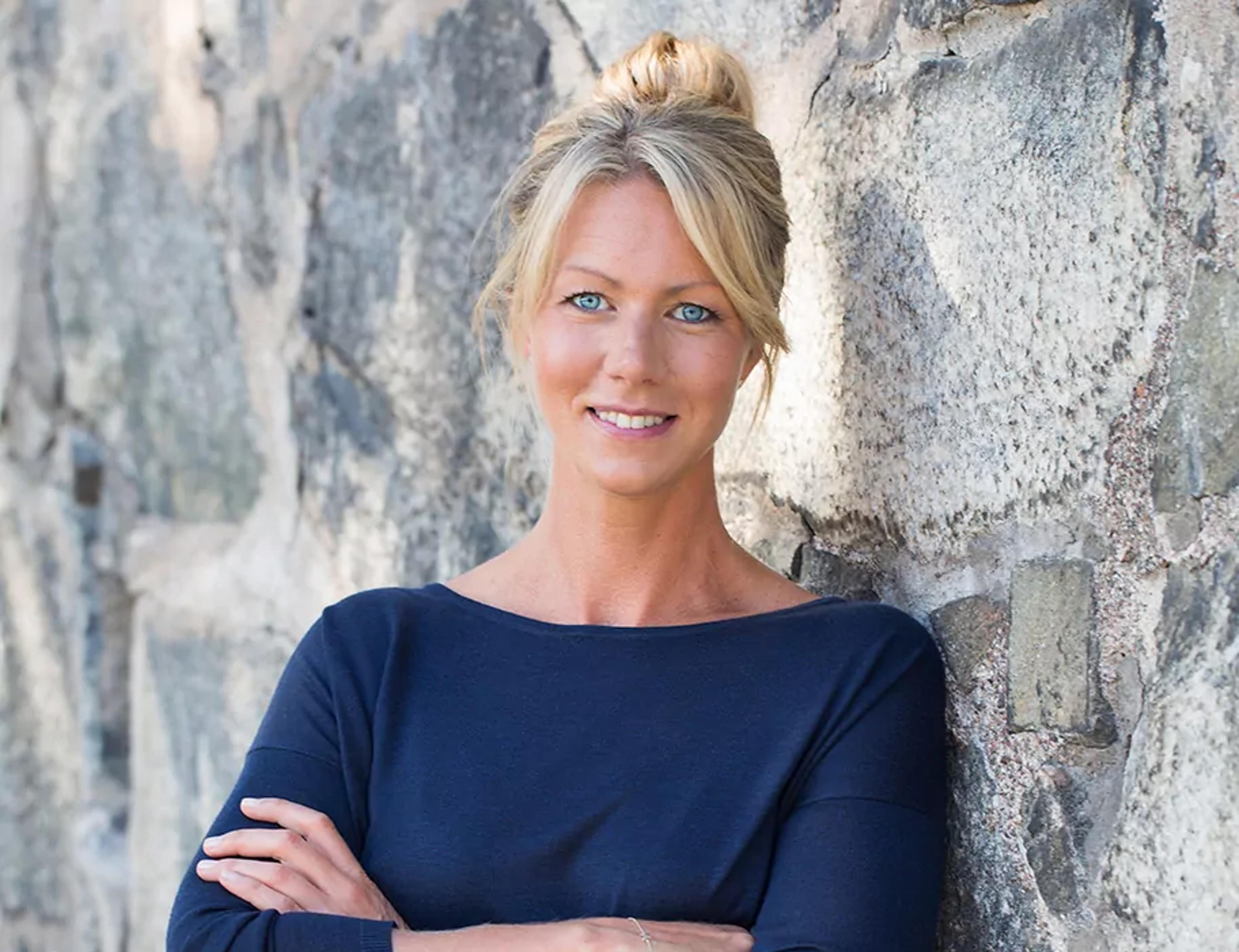 Kvinna med ljusblont hår uppsatt i knut, klädd i en mörkblå tröja, står lutad mot en stenmur med armarna i kors.