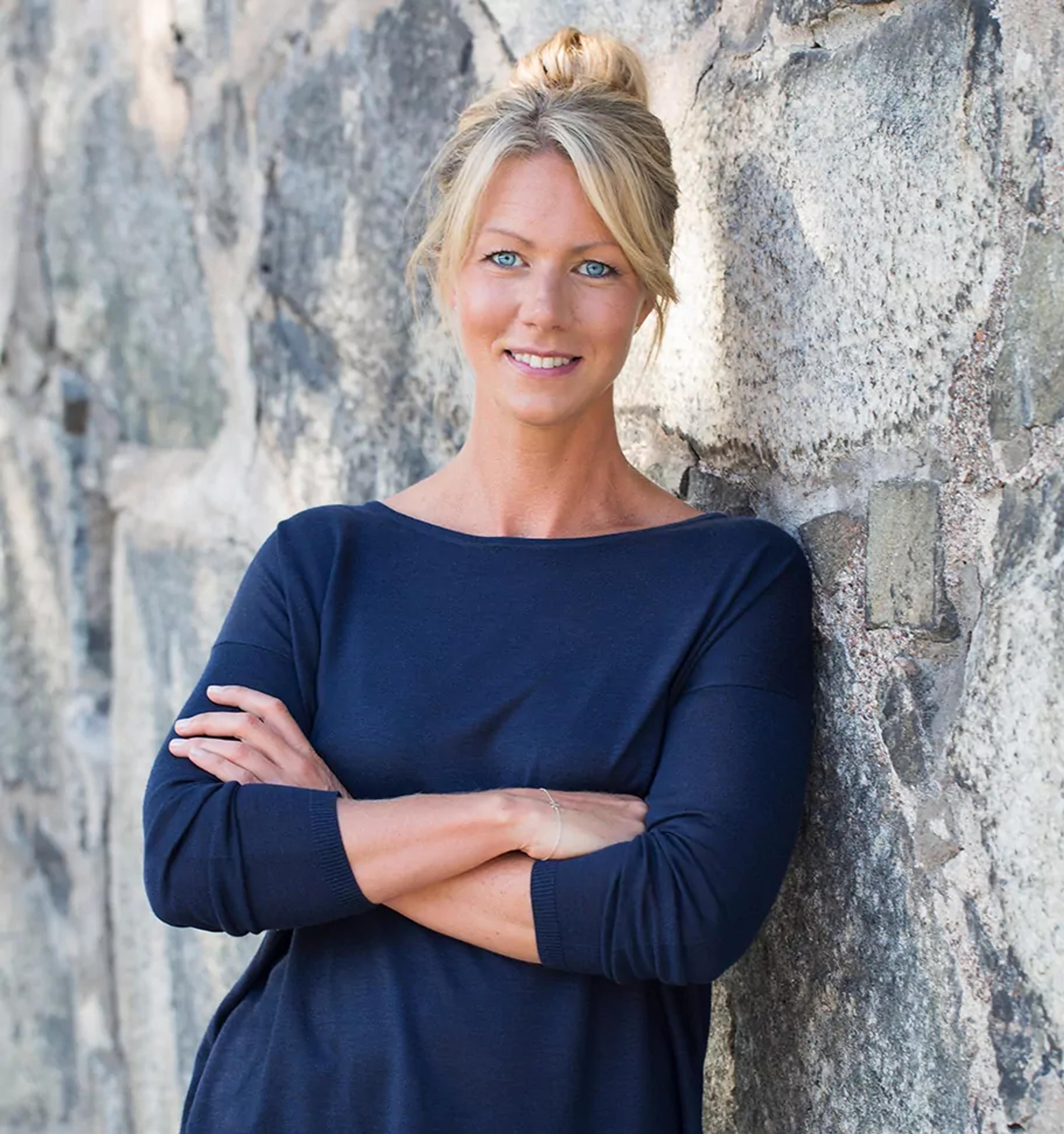 Kvinna med ljusblont hår uppsatt i knut, klädd i en mörkblå tröja, står lutad mot en stenmur med armarna i kors.