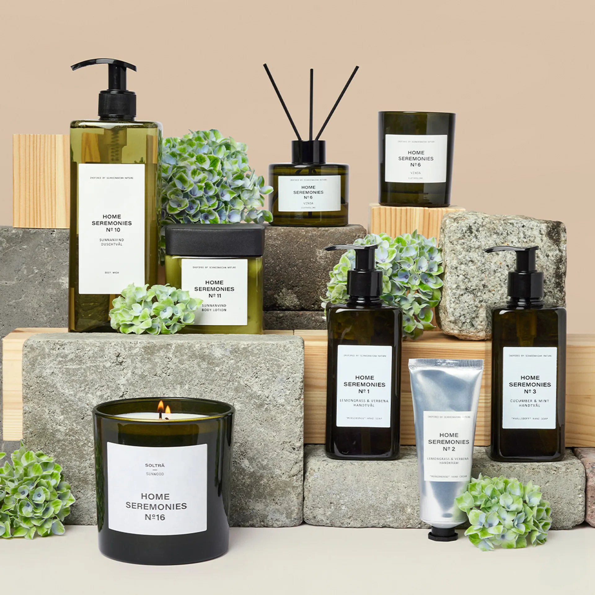 Ett set med doftljus, handtvålar med pump och handkräm i en enkel, stilren design i grönt och vitt. Produkterna är kombinerade med dekorativa gröna blommor mot en bakgrund av betong och träblock.