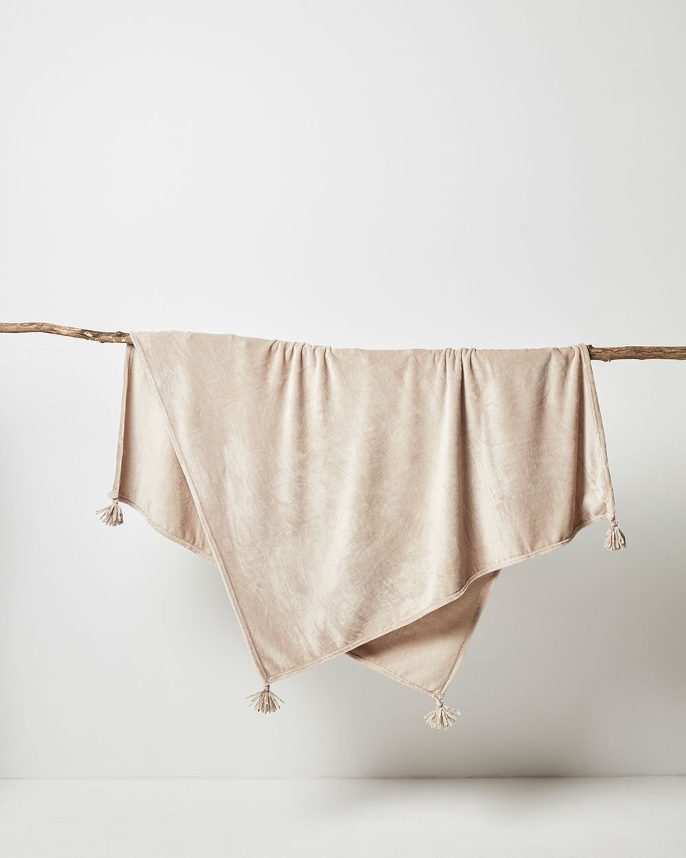 En mjuk, beige fleecefilt med små tofsar i hörnen, hängande över en tunn trägren. Filten har en lätt lyster och en behaglig, varm yta.