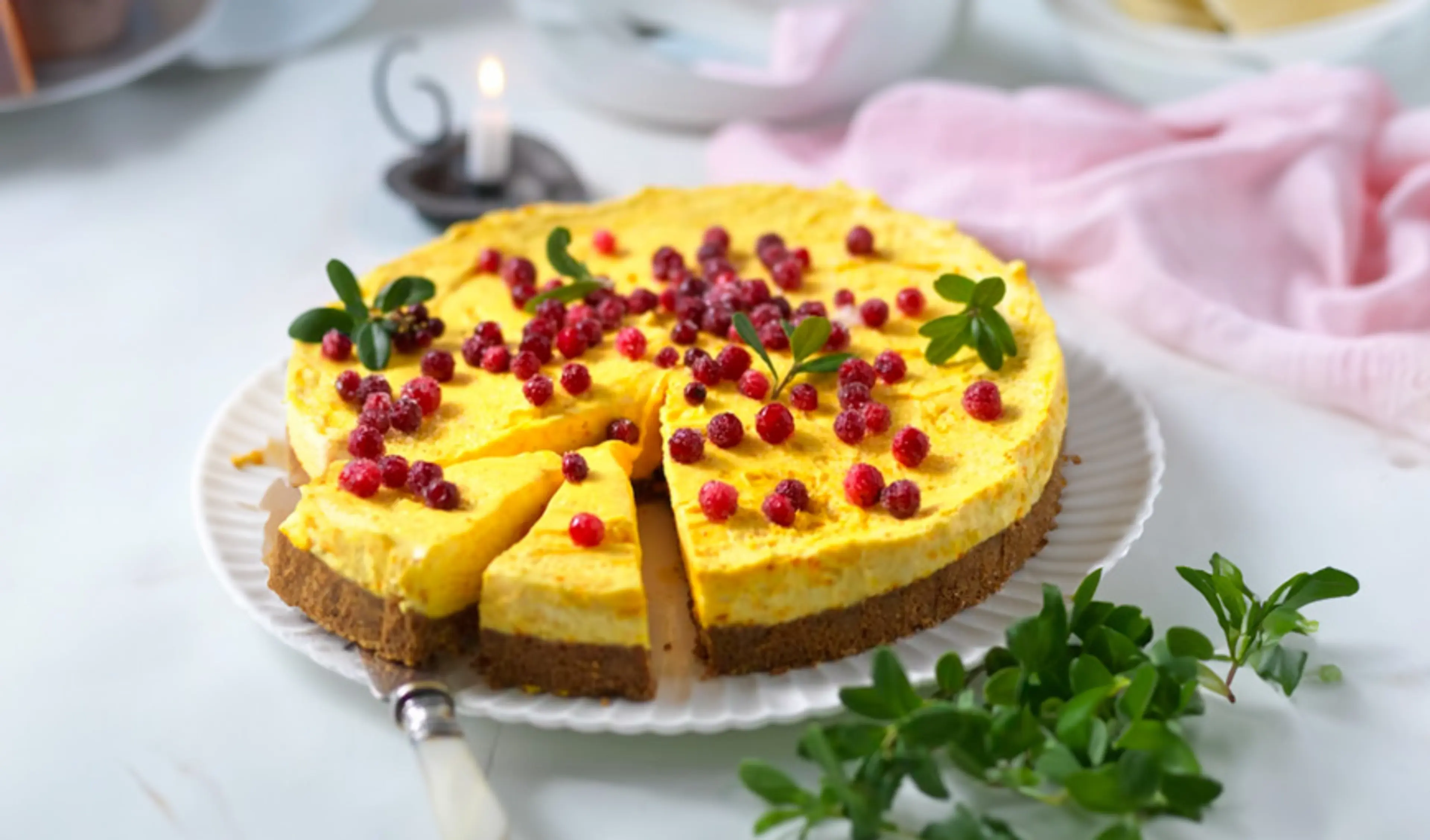 En rund cheesecake med gul fyllning och krispig chokladbotten, garnerad med röda bär och några gröna blad. En bit är redan försiktigt utdragen från kakan.