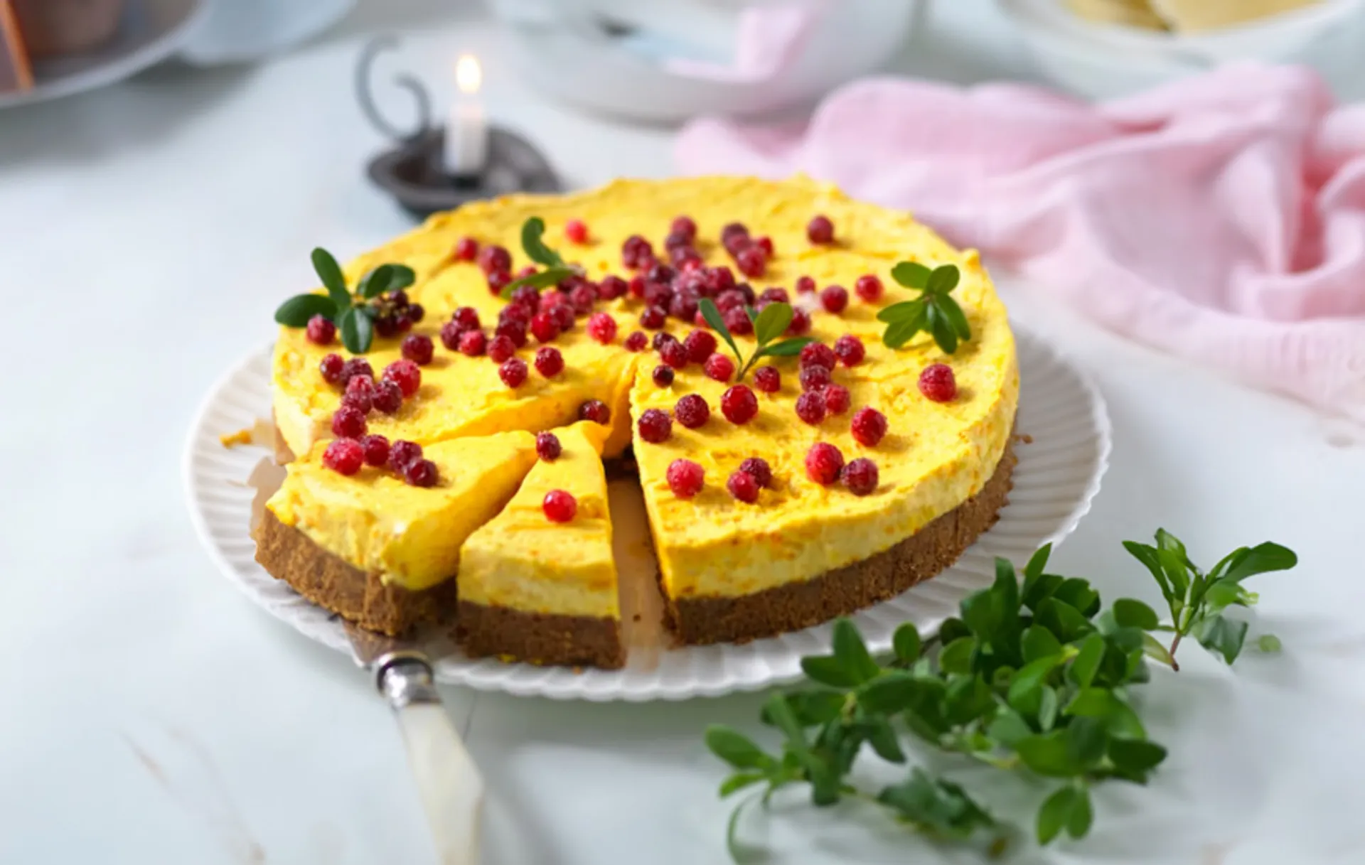 En rund cheesecake med gul fyllning och krispig chokladbotten, garnerad med röda bär och några gröna blad. En bit är redan försiktigt utdragen från kakan.