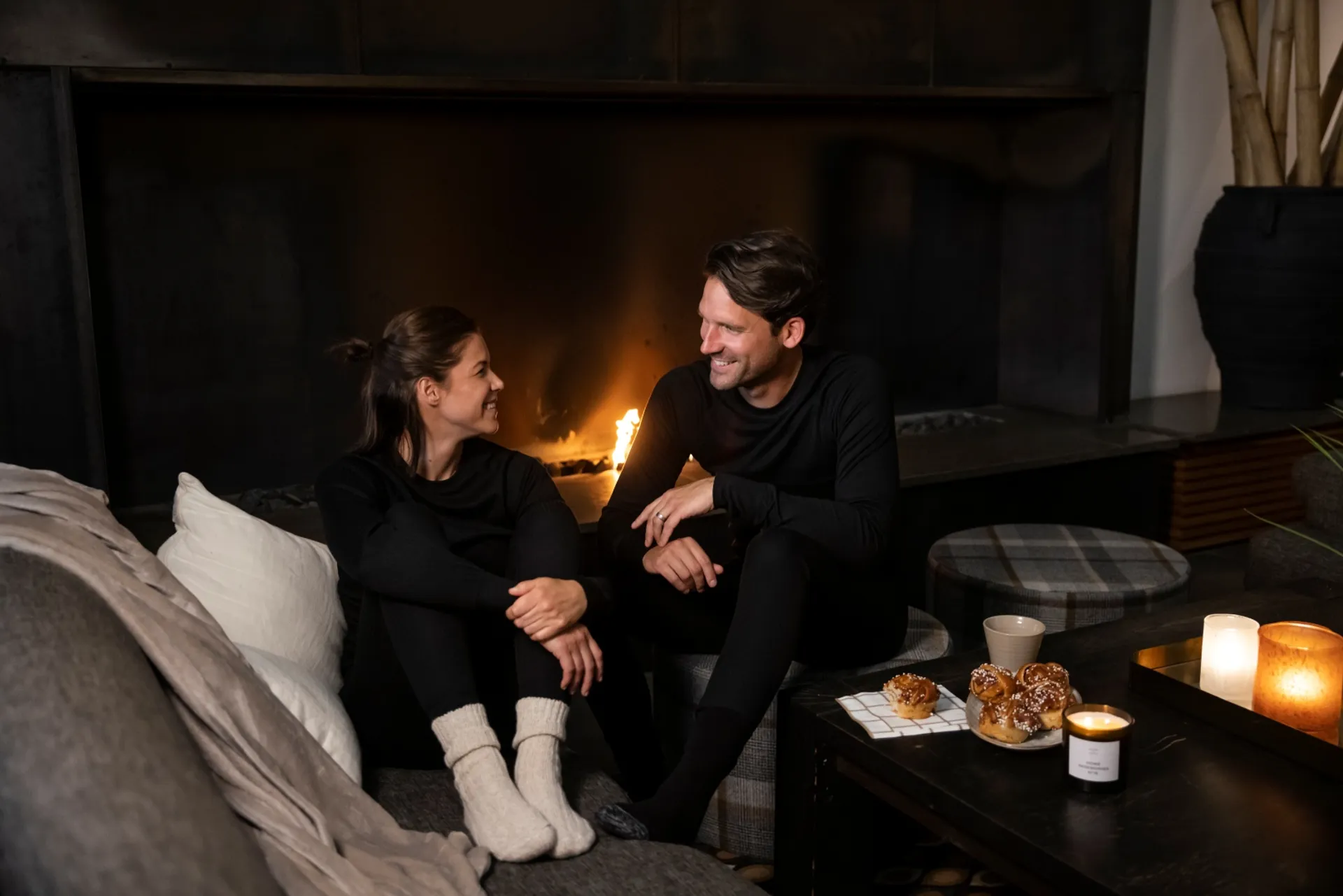 Två personer i svarta, sportiga underställ sitter avslappnat framför en brasa, den ena med varma, stickade sockor. På bordet bredvid ligger nybakade kanelbullar, en mugg och ett tänt doftljus.