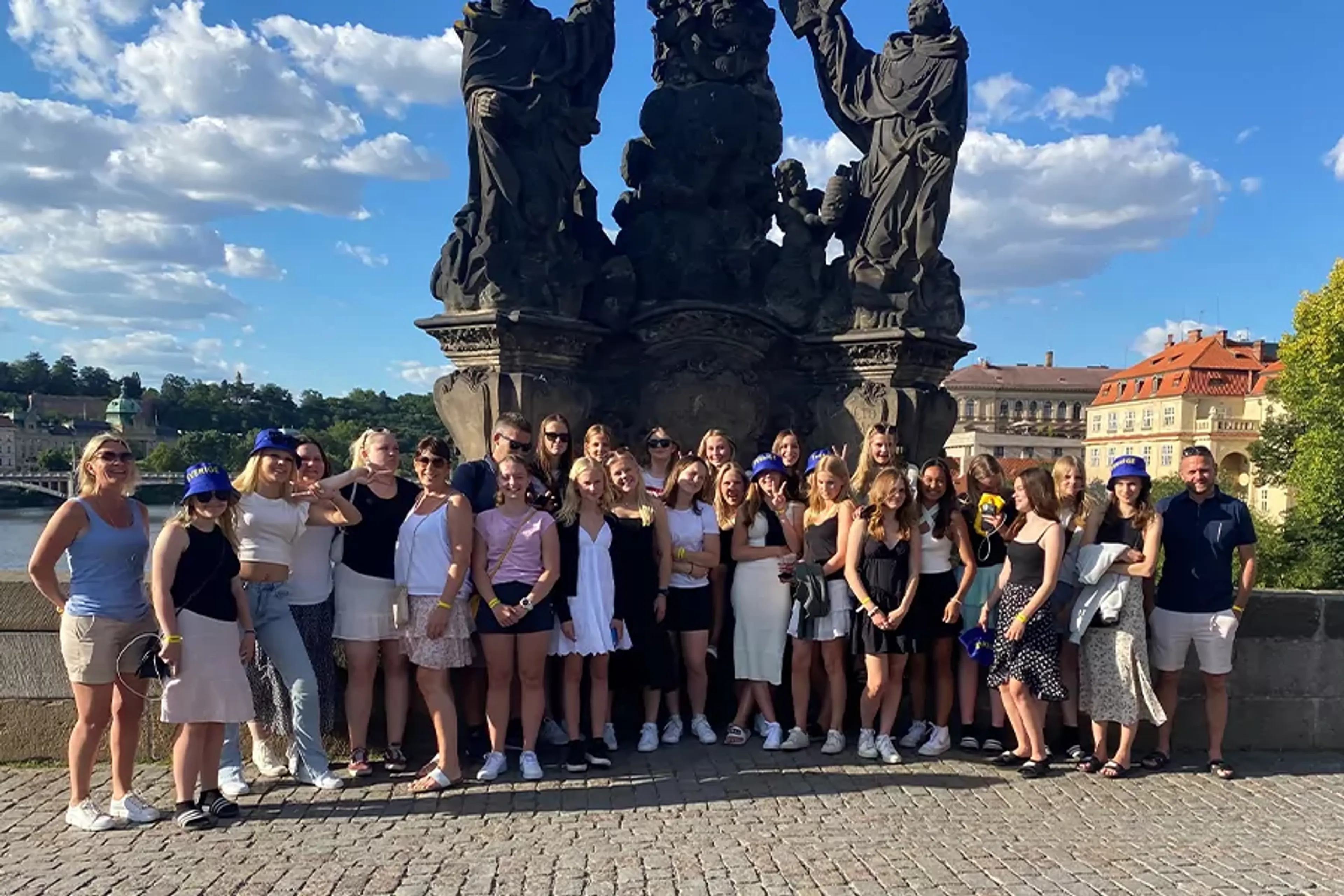 En stor grupp människor står samlade framför en detaljerad stenskulptur på en bro under en solig himmel med moln. I bakgrunden syns vattendrag och historiska byggnader.