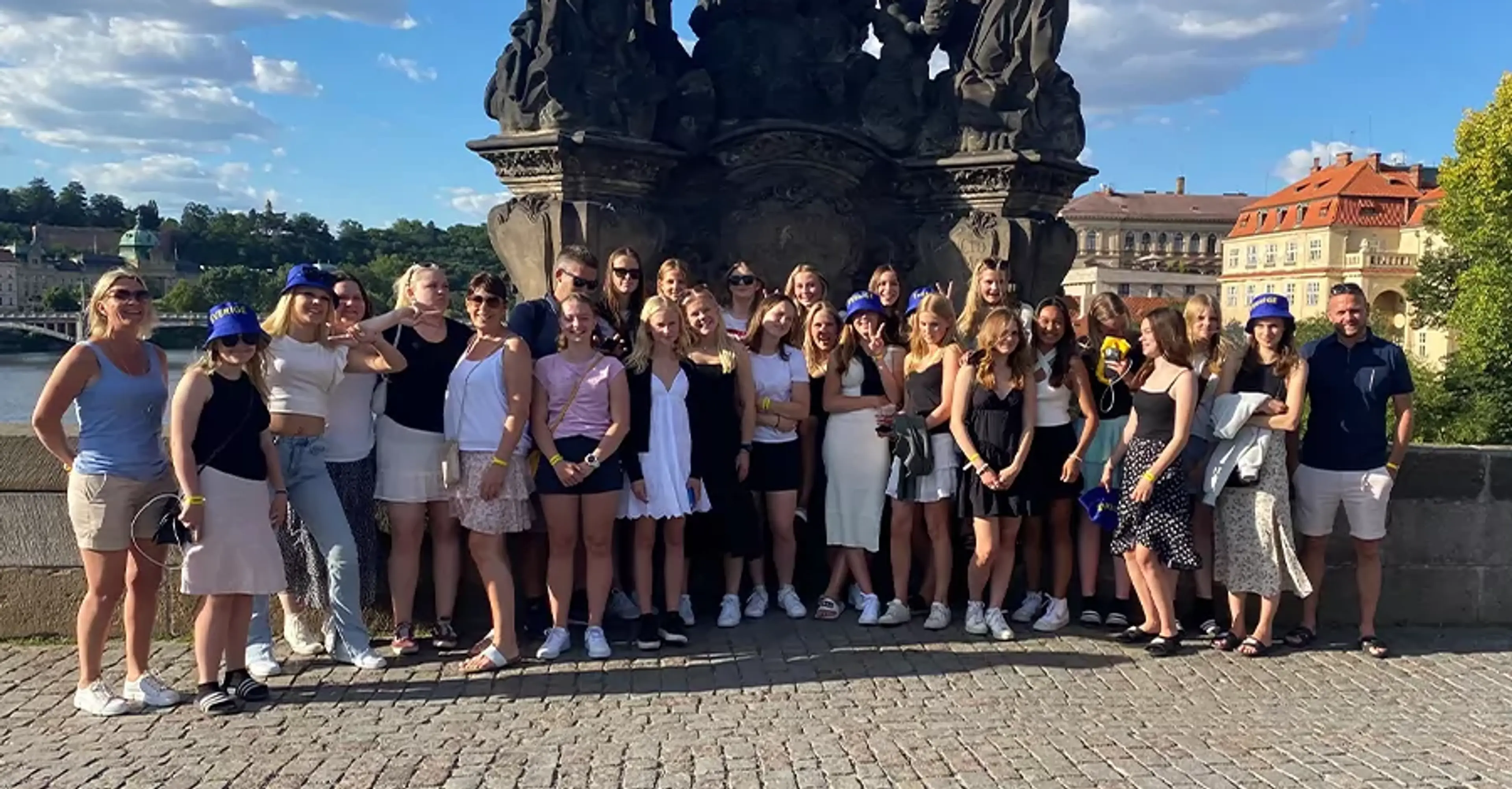 En stor grupp människor står samlade framför en detaljerad stenskulptur på en bro under en solig himmel med moln. I bakgrunden syns vattendrag och historiska byggnader.