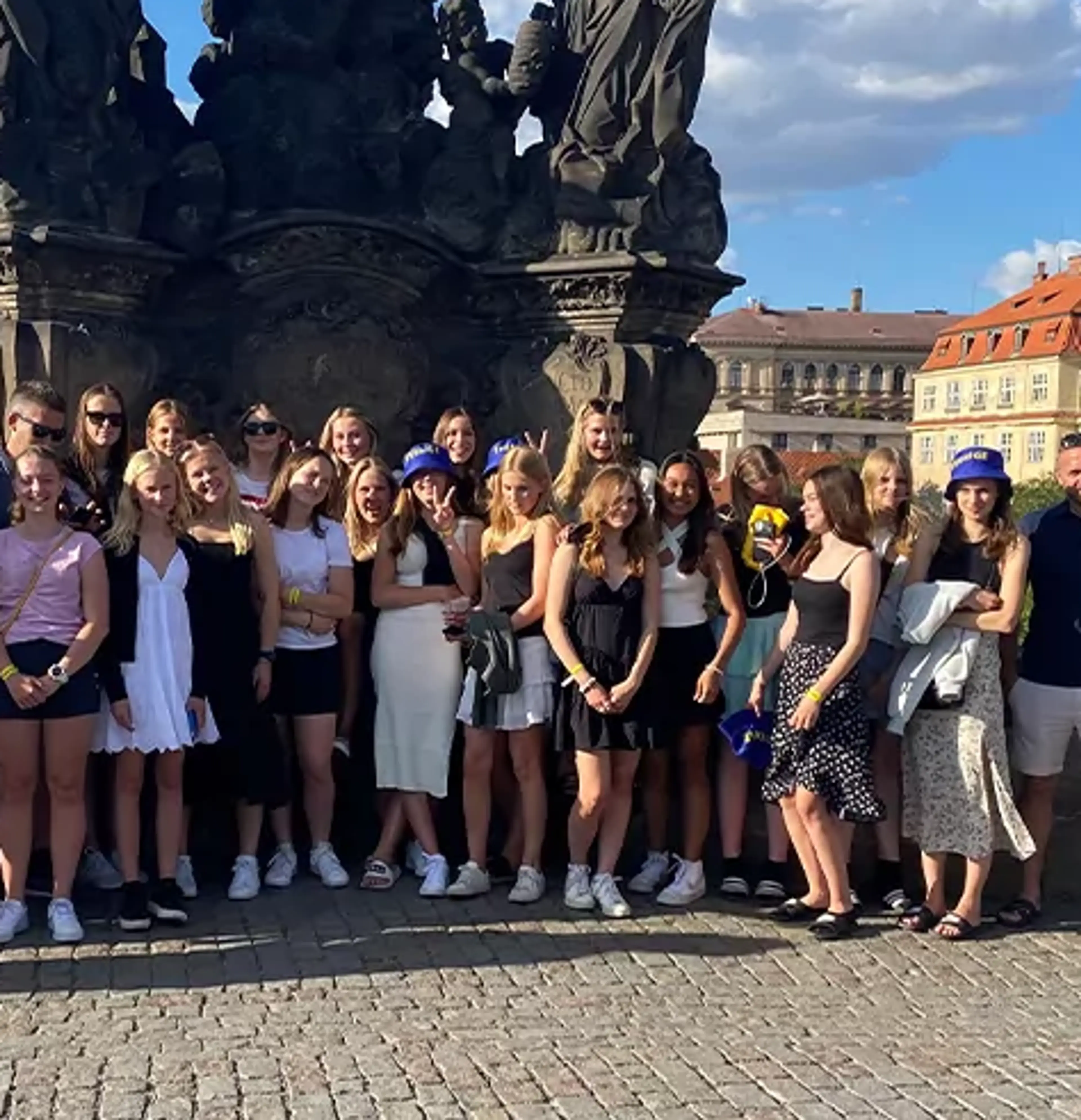 En stor grupp människor står samlade framför en detaljerad stenskulptur på en bro under en solig himmel med moln. I bakgrunden syns vattendrag och historiska byggnader.