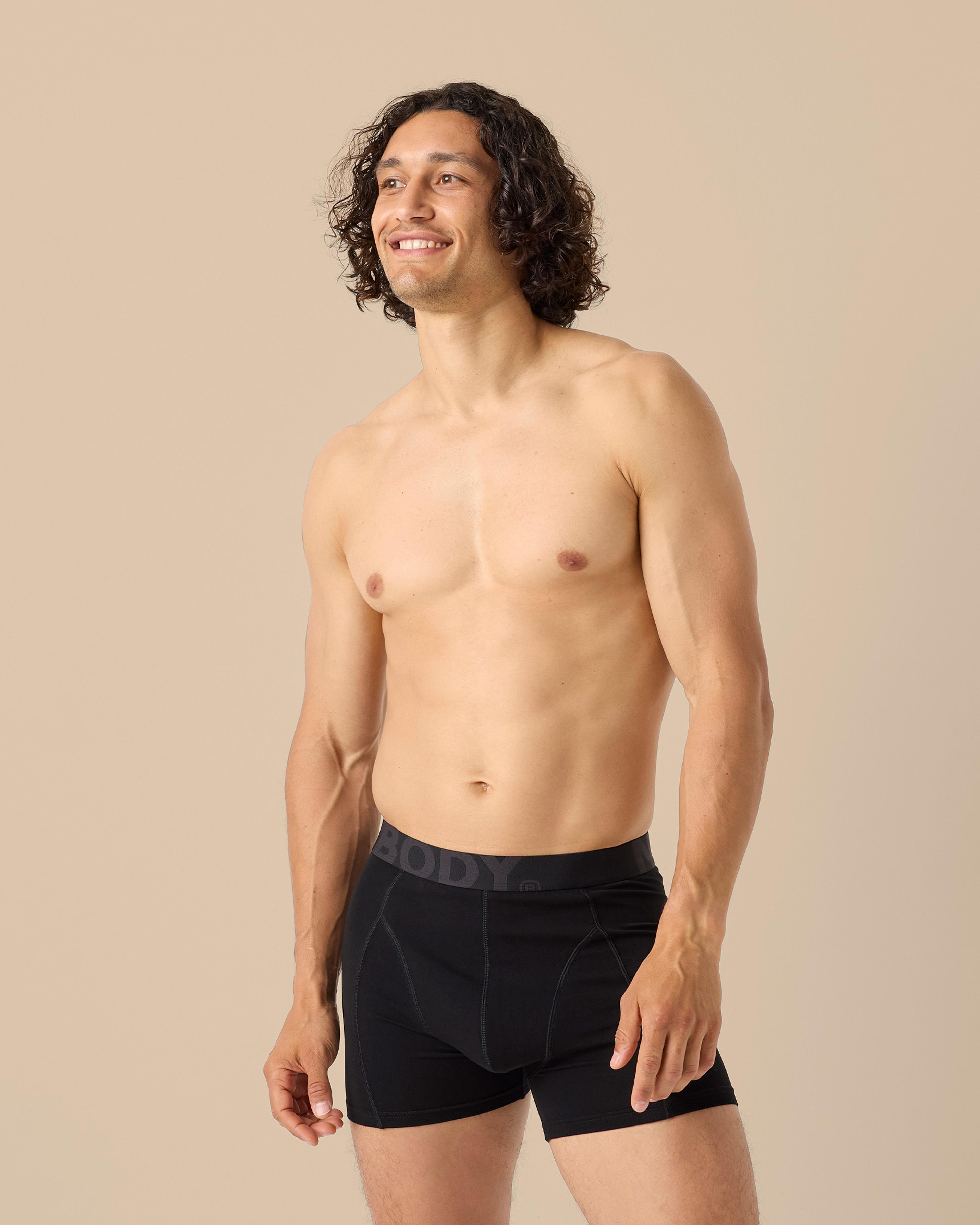 En man bär ett par svarta boxershorts med bred resår i midjan mot en neutral bakgrund. Shortsens sömmar och passform syns tydligt.