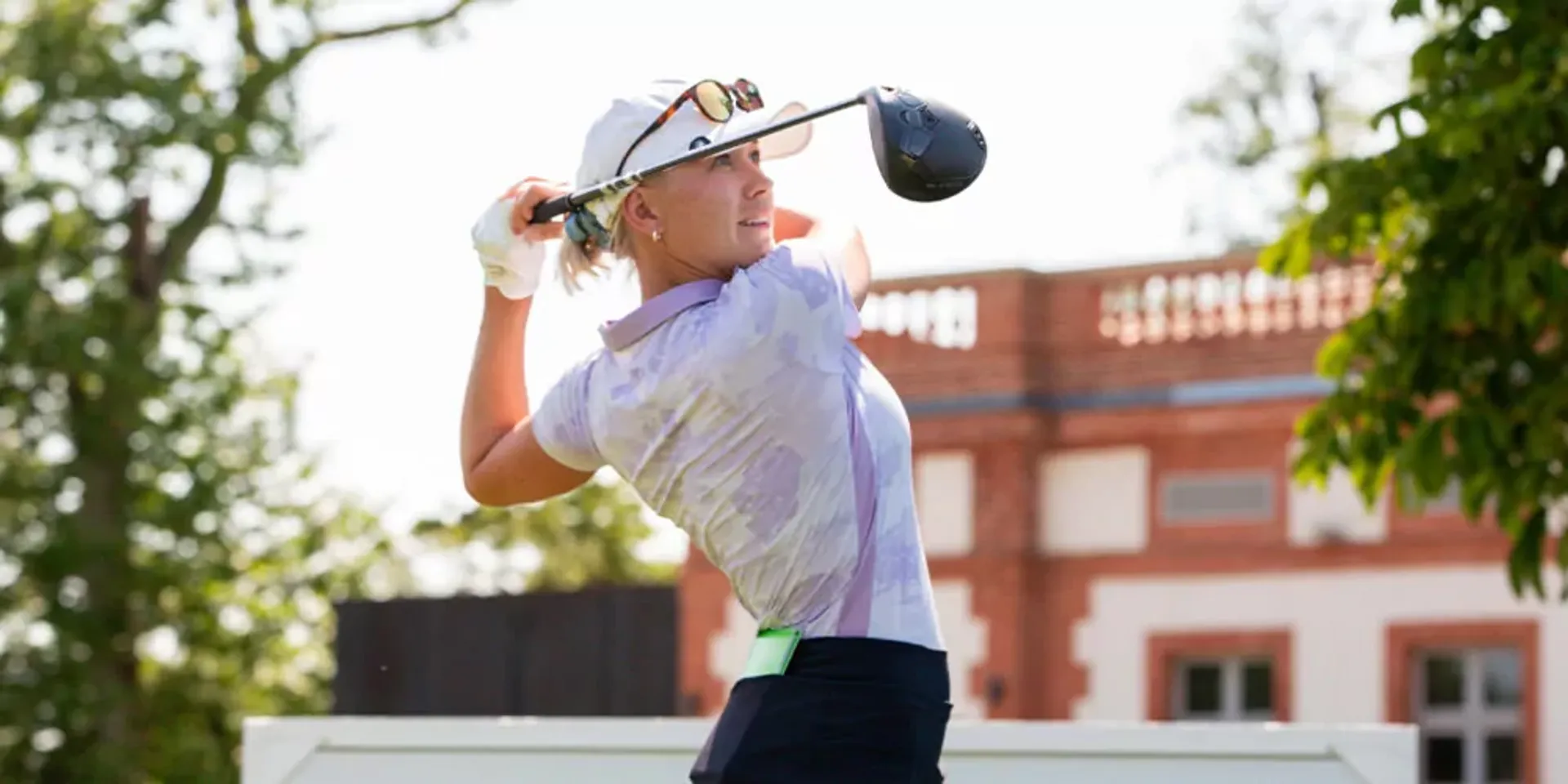 En kvinna i vit keps och ljuslila pikékläder svingar en golfklubba utomhus med en byggnad och grönska i bakgrunden.