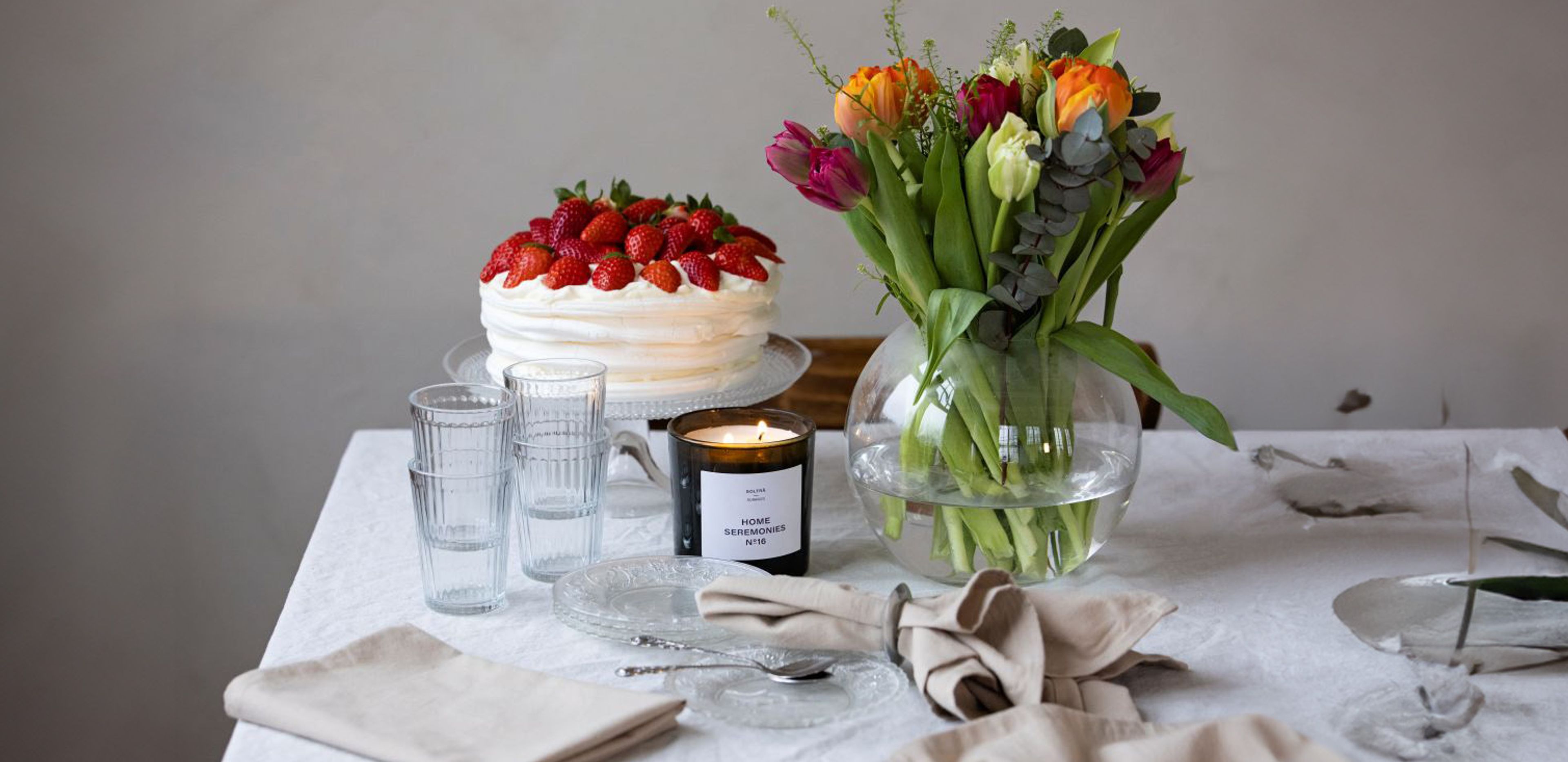 Ett svart doftljus märkt "Home Seremonies" brinner bredvid en rund glasvas med gröna stjälkar, omgivna av klara glas och en vacker glasskål på ett ljust dukat bord.