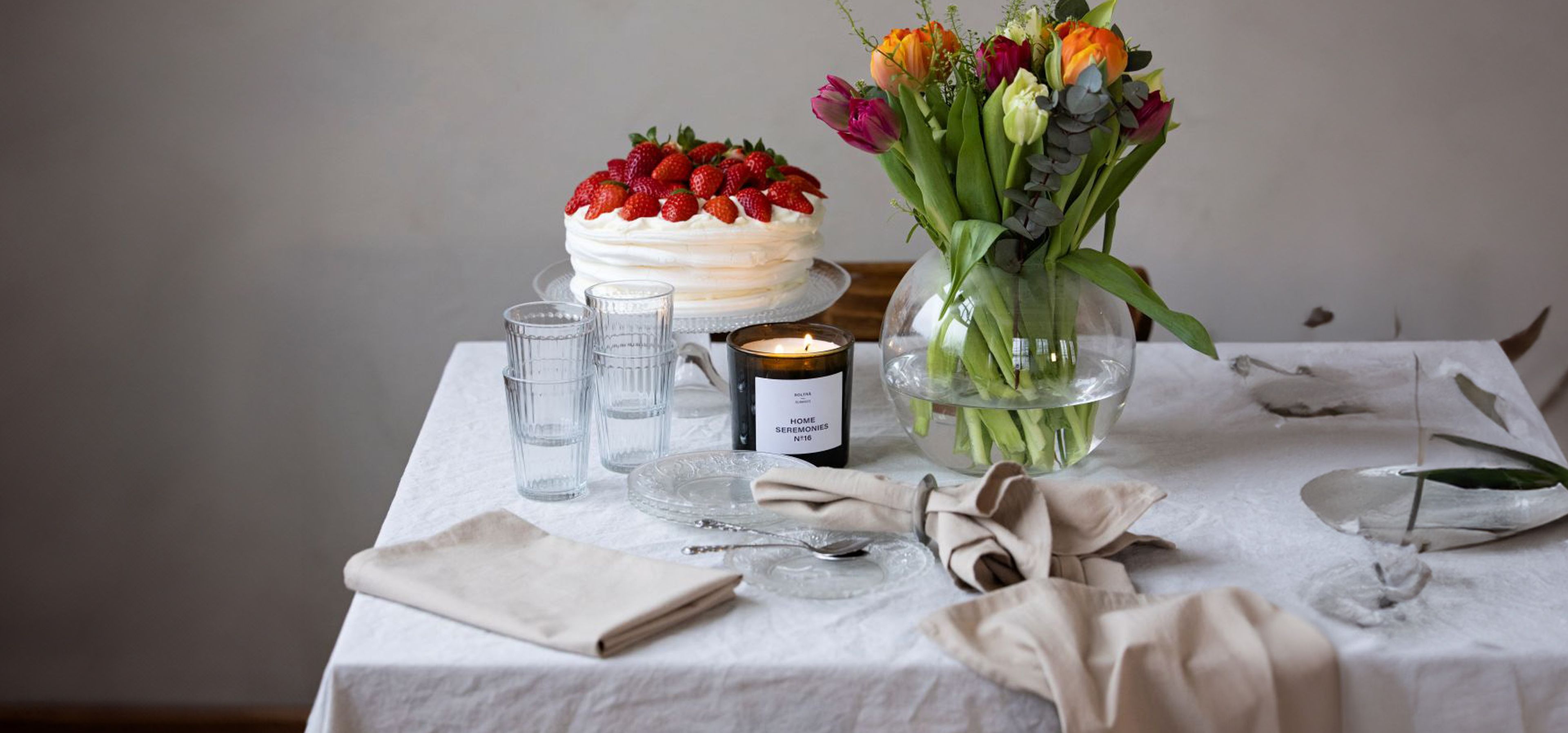 Ett svart doftljus märkt "Home Seremonies" brinner bredvid en rund glasvas med gröna stjälkar, omgivna av klara glas och en vacker glasskål på ett ljust dukat bord.