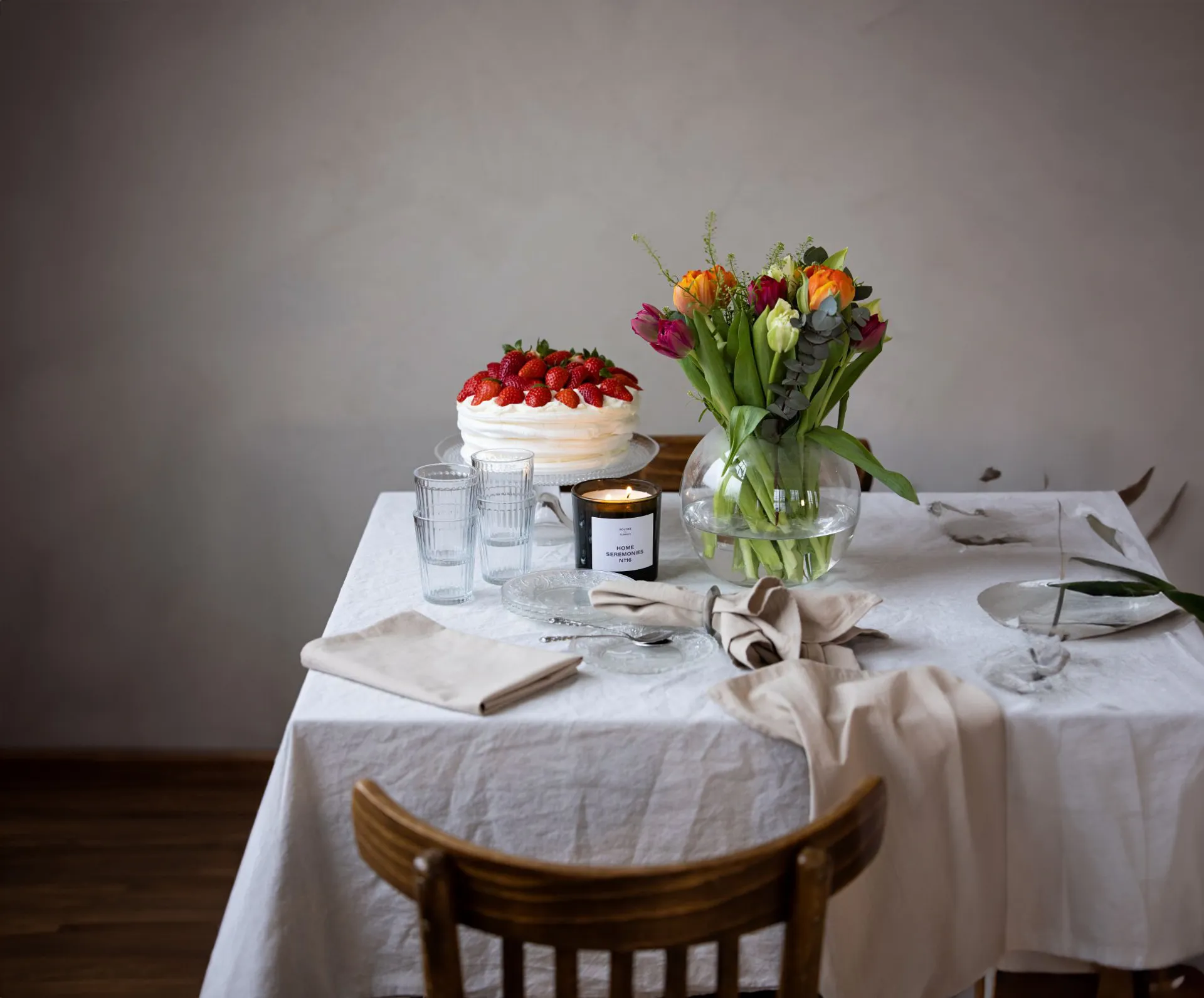 Ett svart doftljus märkt "Home Seremonies" brinner bredvid en rund glasvas med gröna stjälkar, omgivna av klara glas och en vacker glasskål på ett ljust dukat bord.