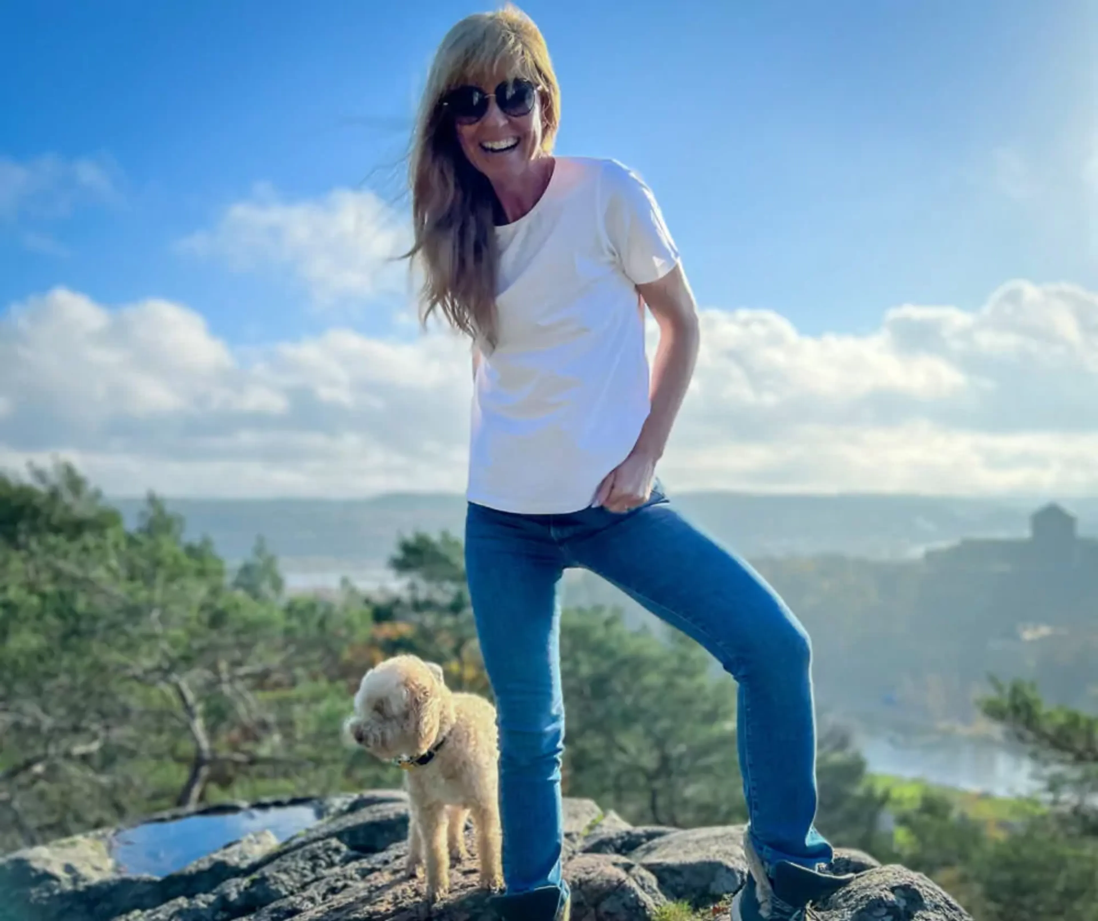 En kvinna i en vit t-shirt och blå jeans står på en klippa tillsammans med en liten beige hund, med en vidsträckt natur och blå himmel i bakgrunden.