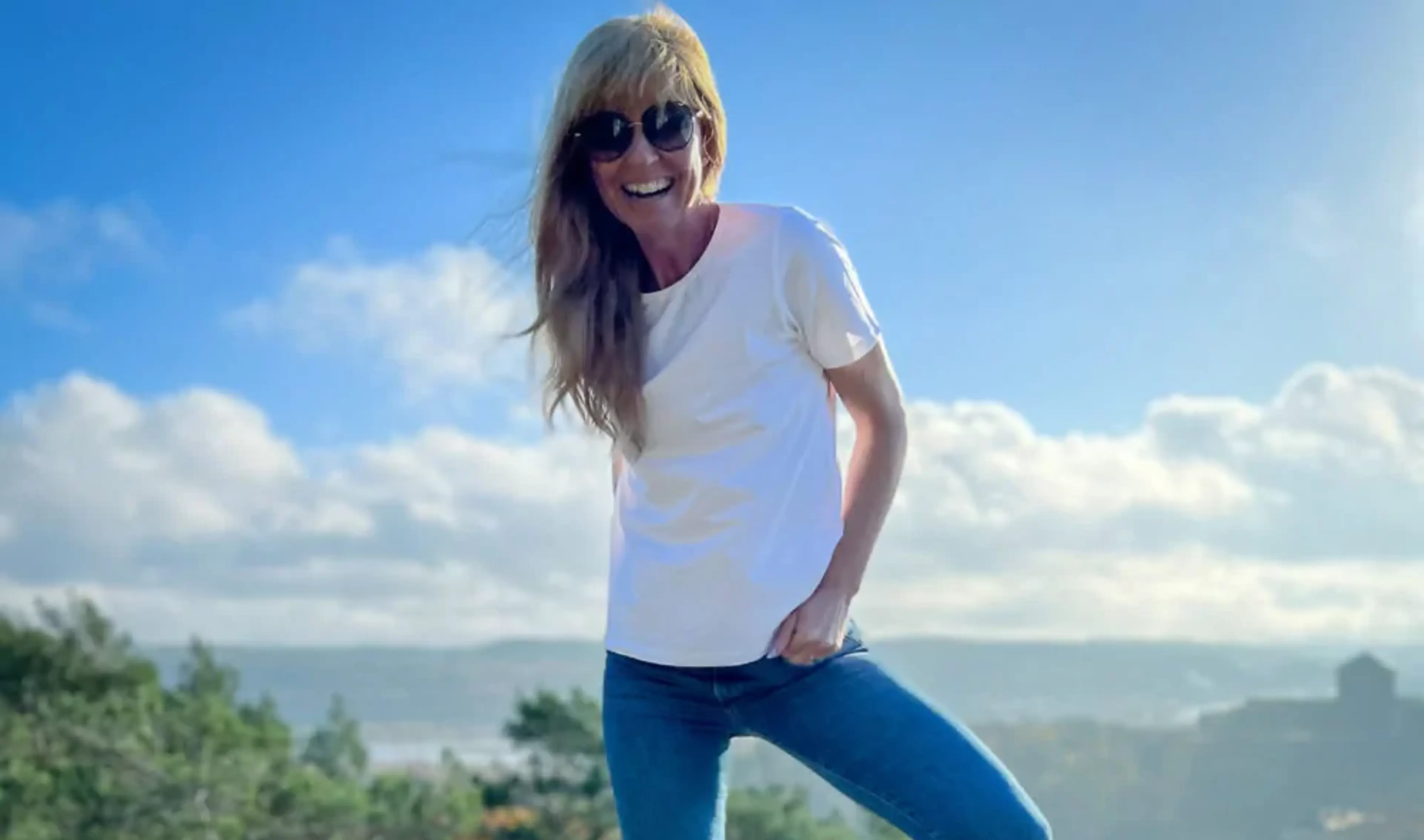 En kvinna i en vit t-shirt och blå jeans står på en klippa tillsammans med en liten beige hund, med en vidsträckt natur och blå himmel i bakgrunden.