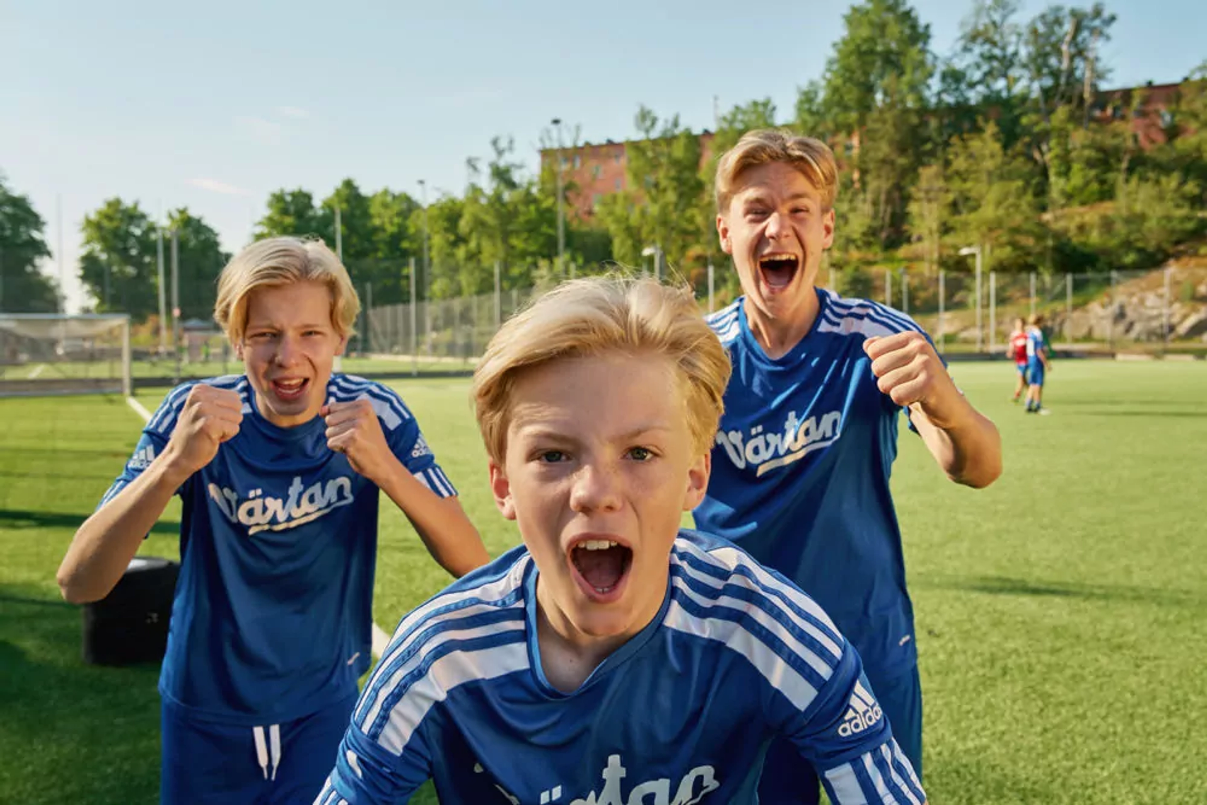 Tre pojkar i blå fotbollströjor står på en gräsplan och uttrycker glädje och pepp. Bakgrunden visar en solig dag med träd och byggnader.