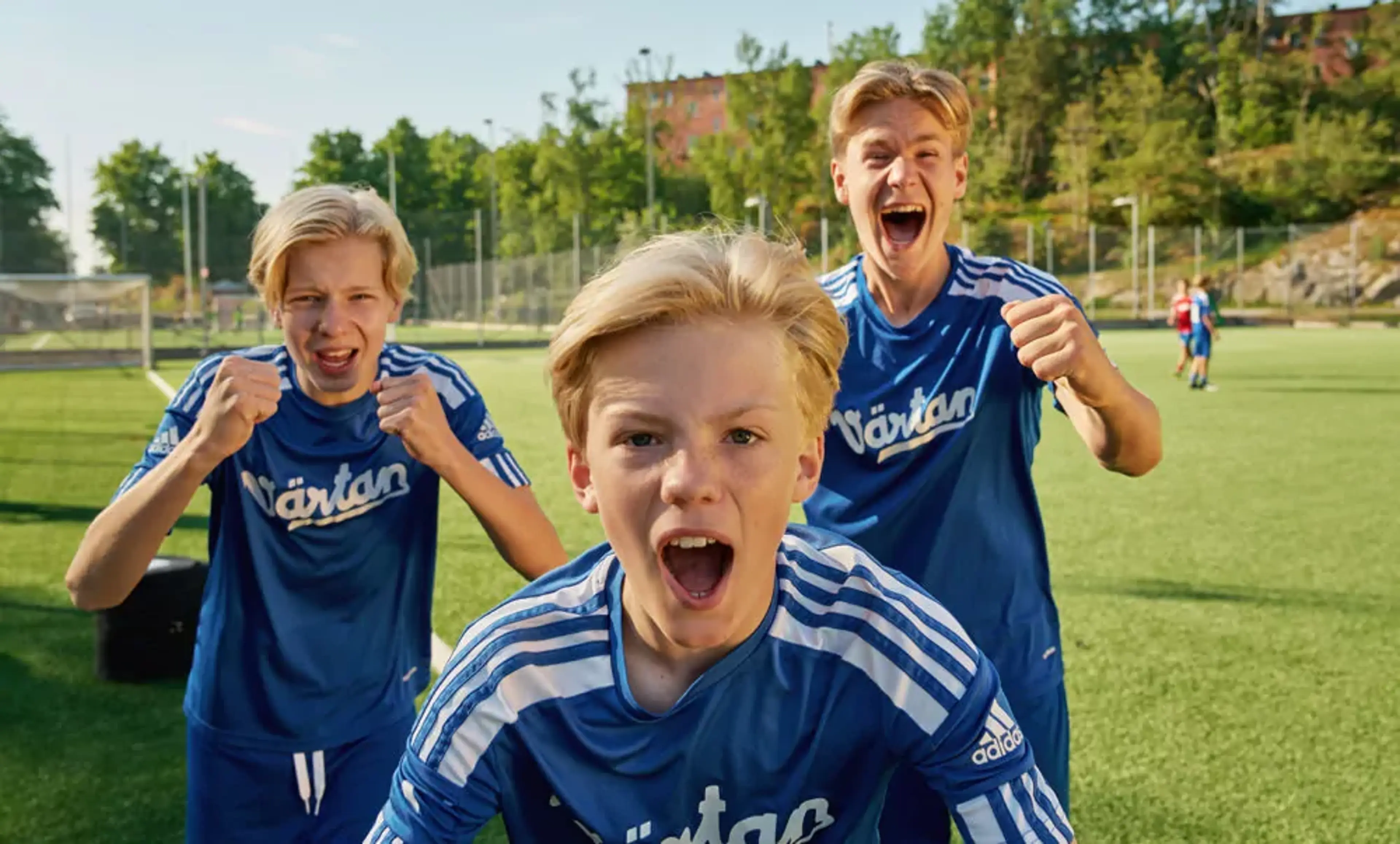 Tre pojkar i blå fotbollströjor står på en gräsplan och uttrycker glädje och pepp. Bakgrunden visar en solig dag med träd och byggnader.
