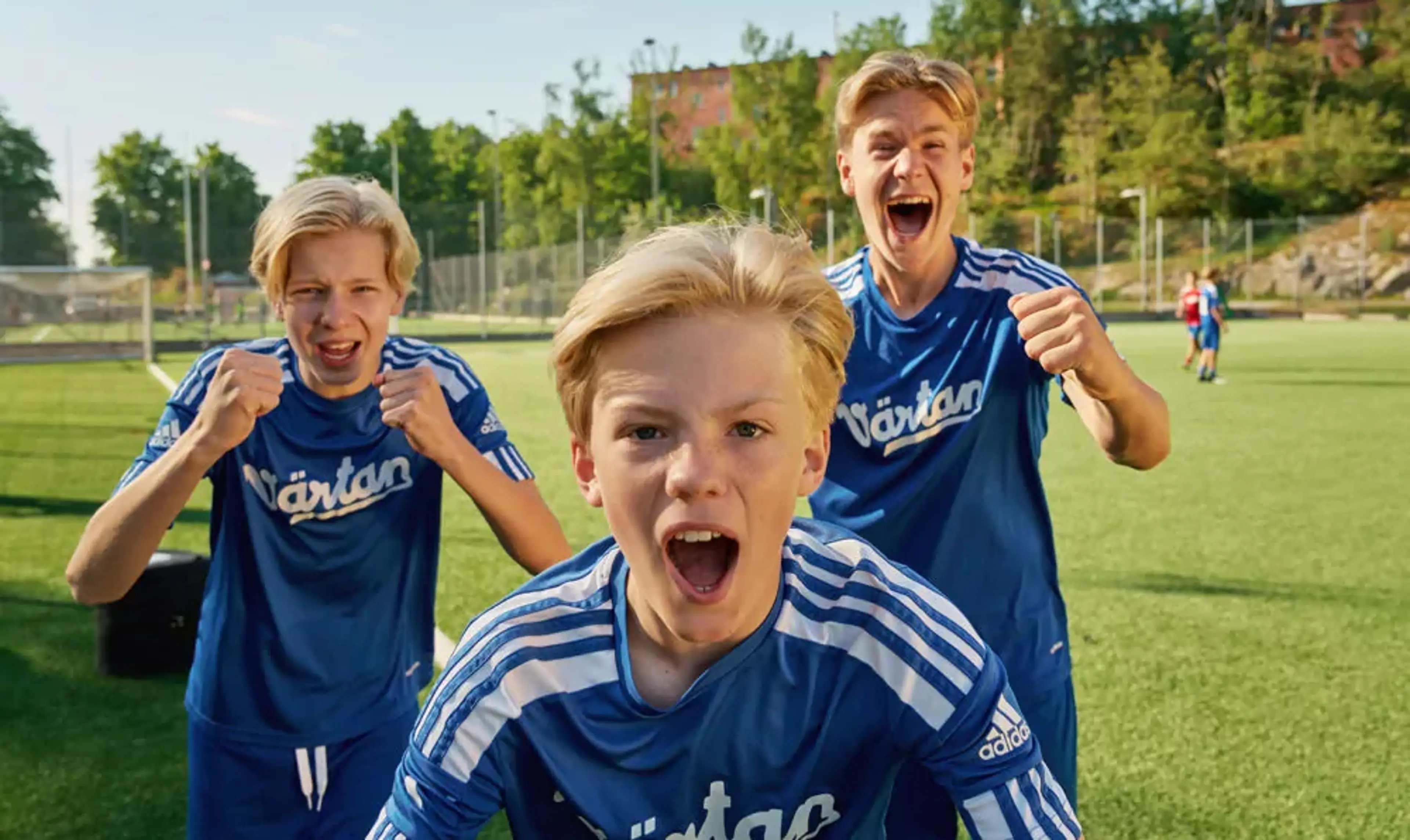 Tre pojkar i blå fotbollströjor står på en gräsplan och uttrycker glädje och pepp. Bakgrunden visar en solig dag med träd och byggnader.