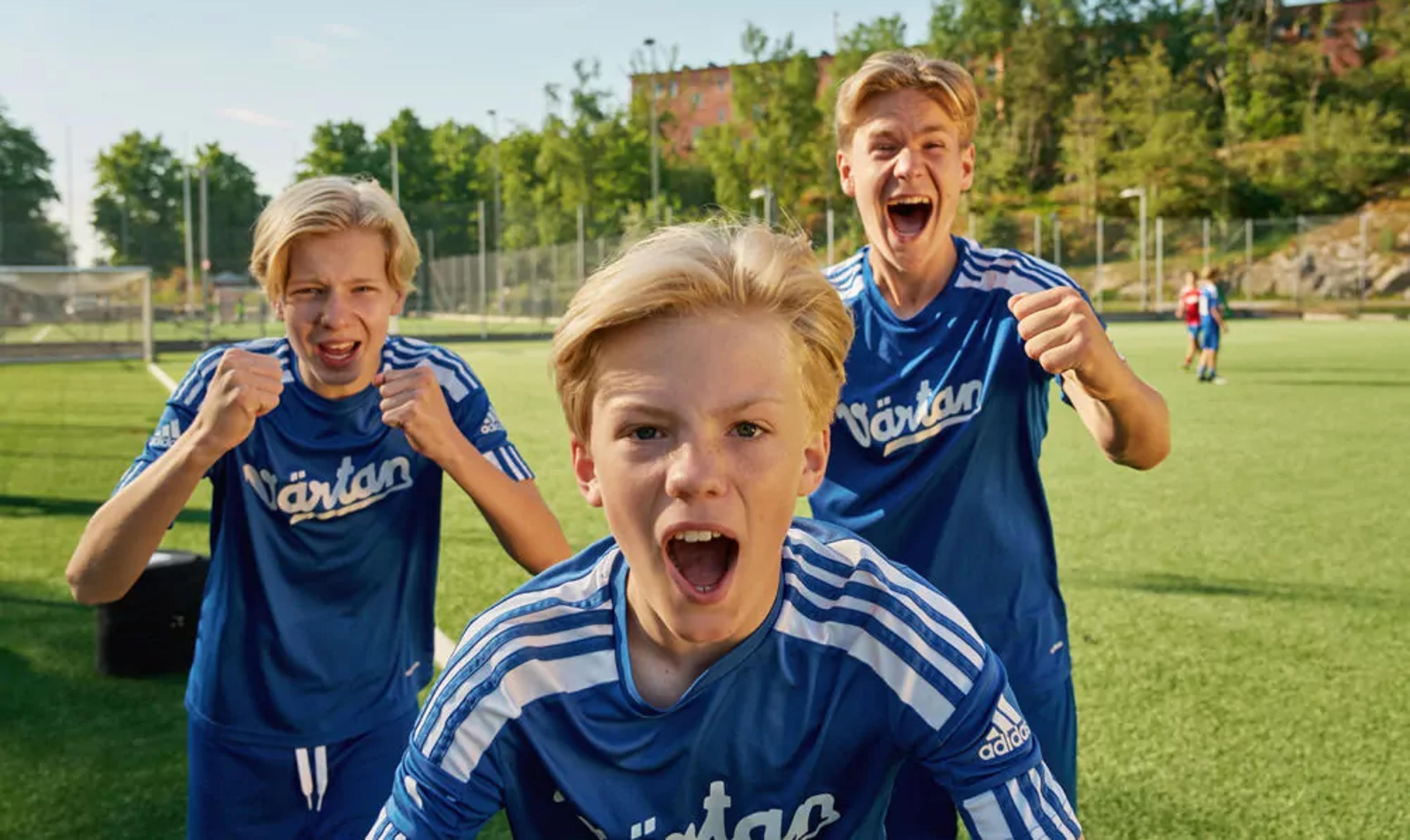 Tre pojkar i blå fotbollströjor står på en gräsplan och uttrycker glädje och pepp. Bakgrunden visar en solig dag med träd och byggnader.