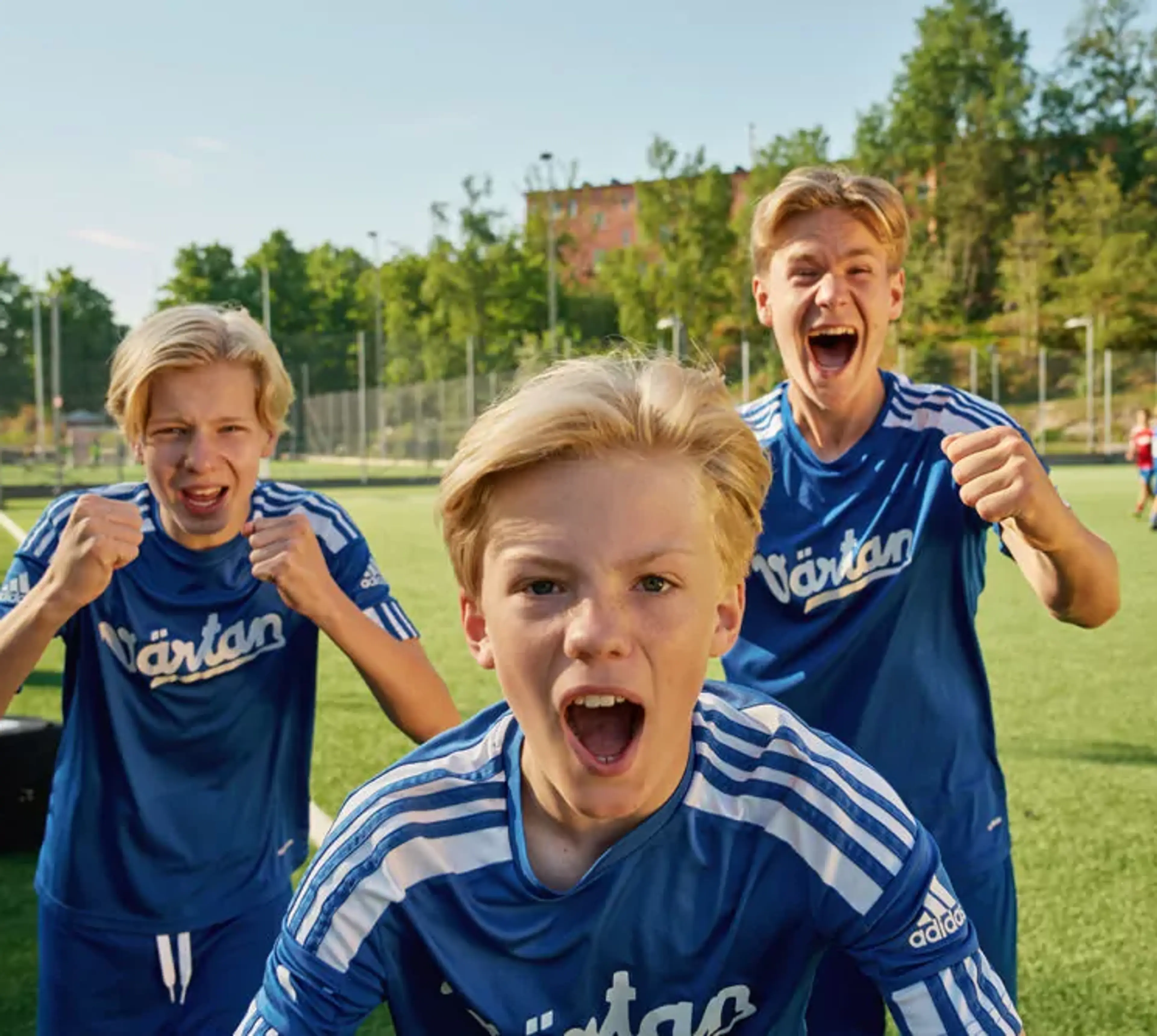 Tre pojkar i blå fotbollströjor står på en gräsplan och uttrycker glädje och pepp. Bakgrunden visar en solig dag med träd och byggnader.