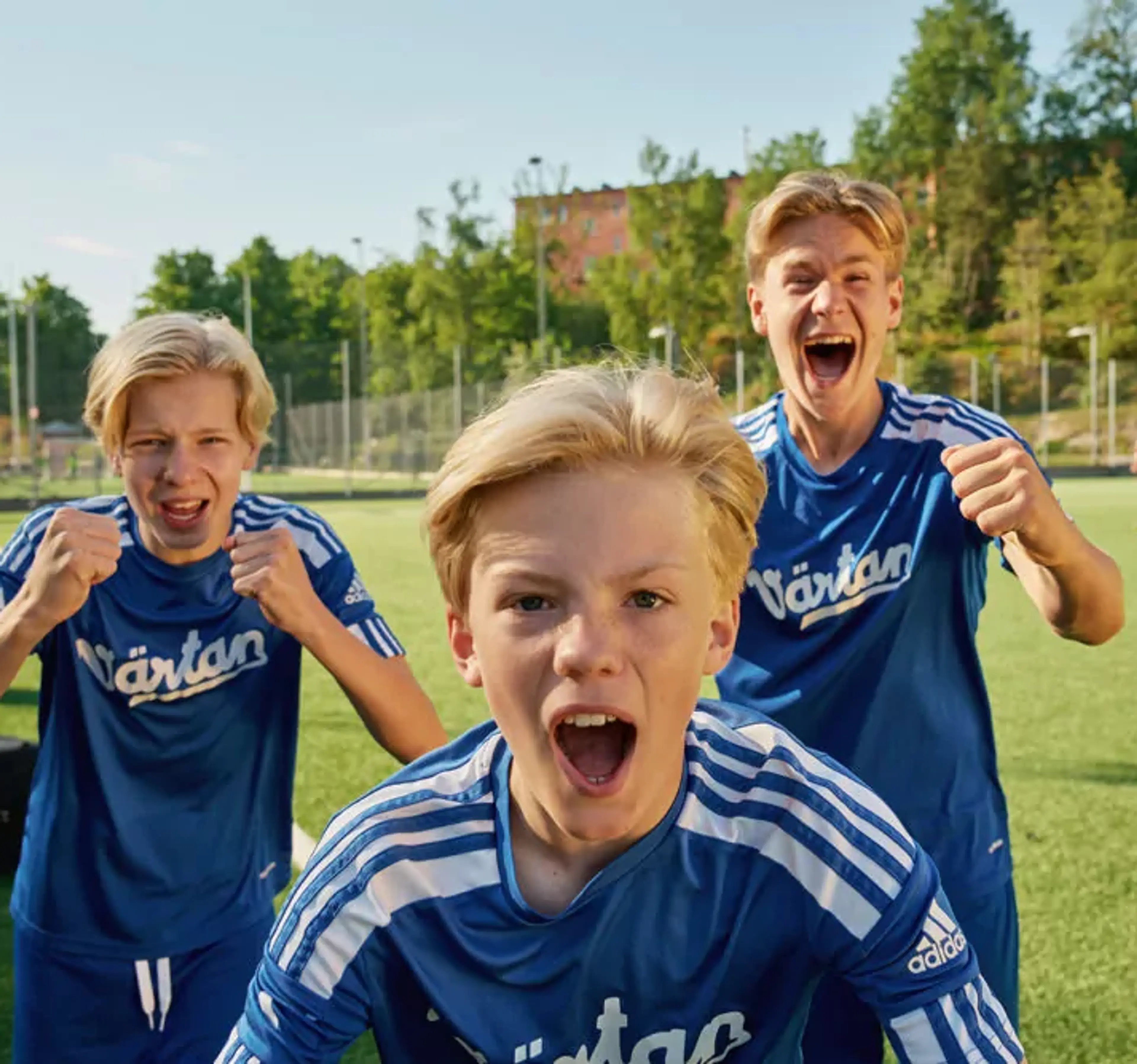 Tre pojkar i blå fotbollströjor står på en gräsplan och uttrycker glädje och pepp. Bakgrunden visar en solig dag med träd och byggnader.