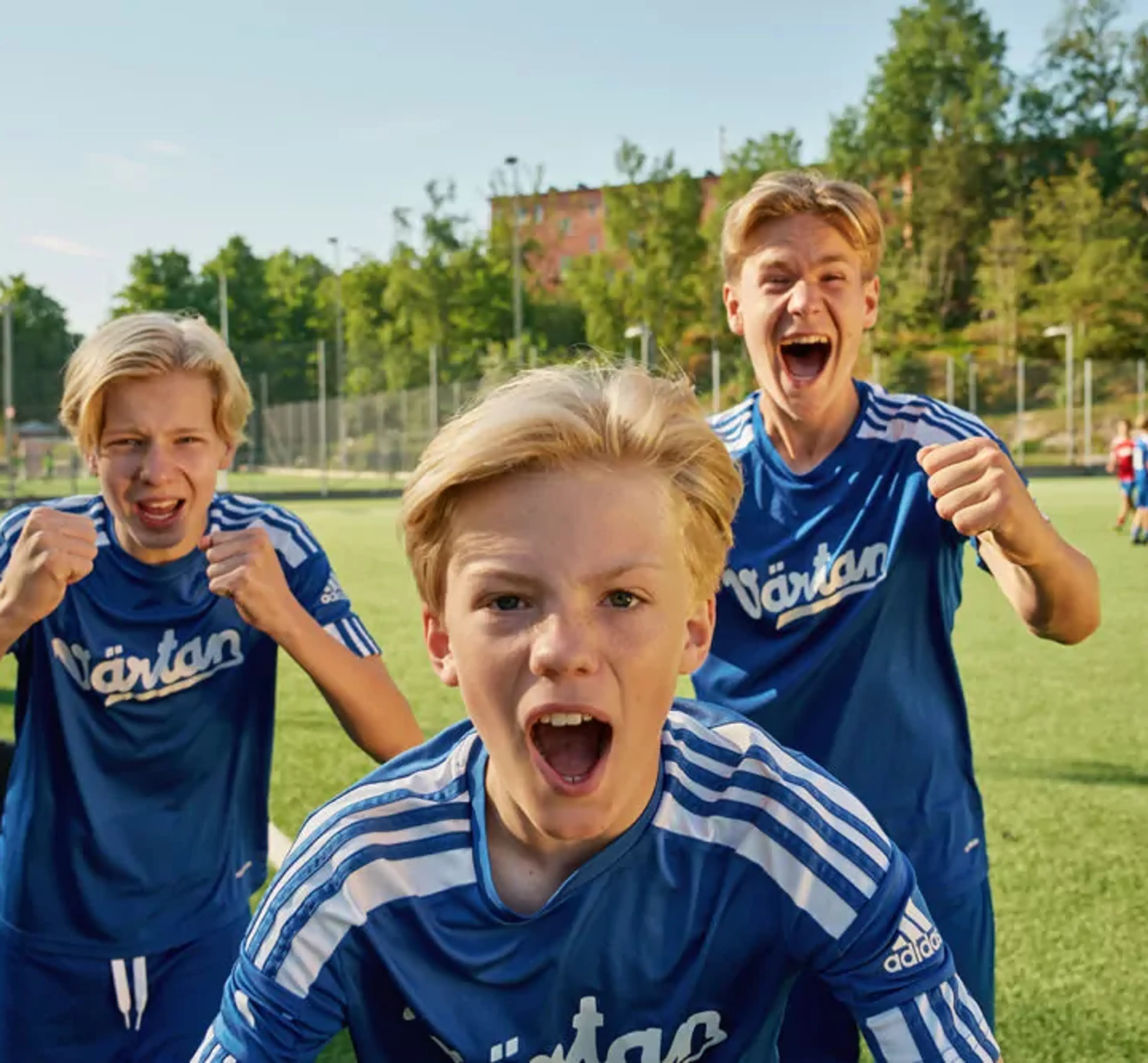 Tre pojkar i blå fotbollströjor står på en gräsplan och uttrycker glädje och pepp. Bakgrunden visar en solig dag med träd och byggnader.