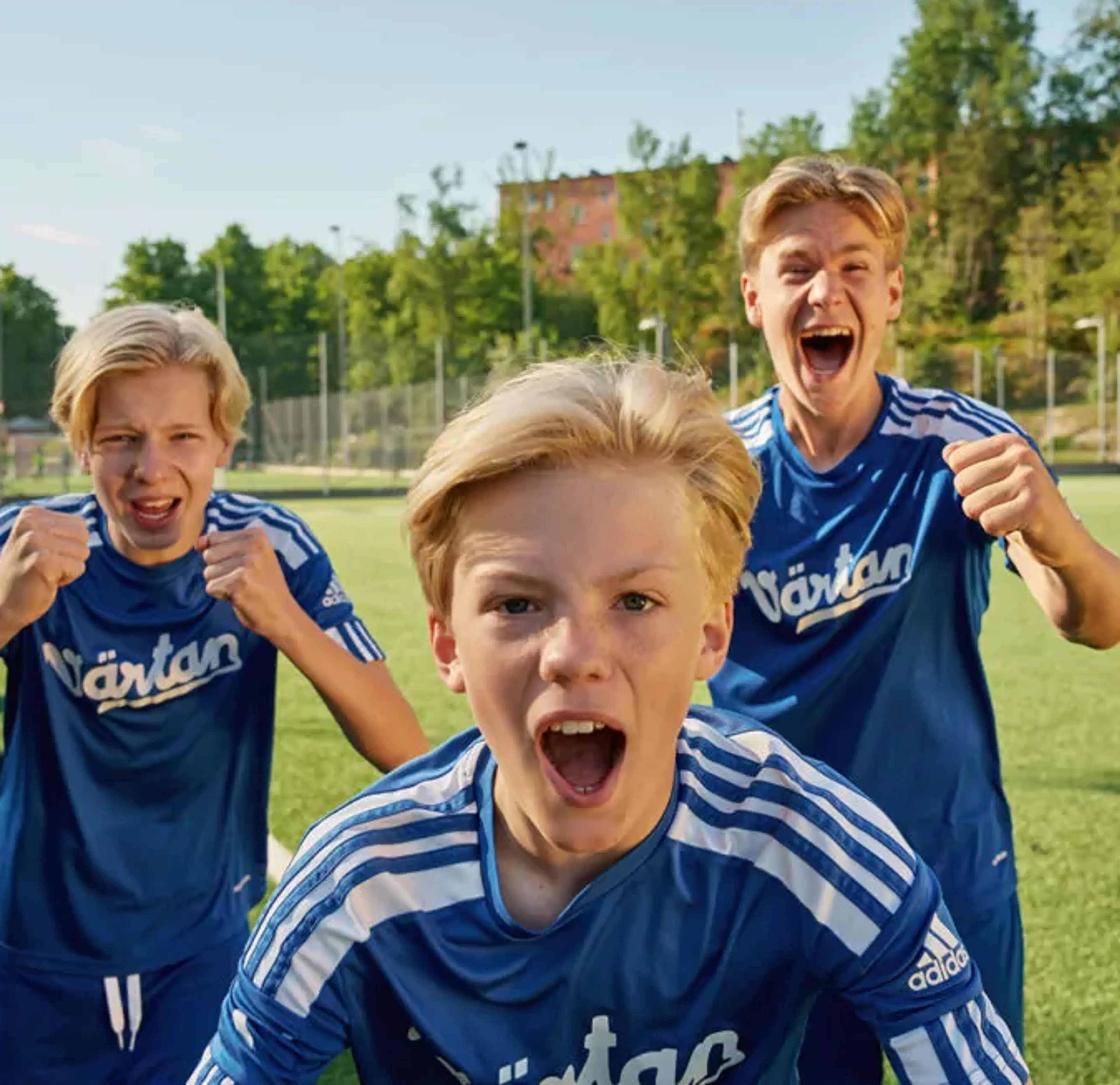Tre pojkar i blå fotbollströjor står på en gräsplan och uttrycker glädje och pepp. Bakgrunden visar en solig dag med träd och byggnader.