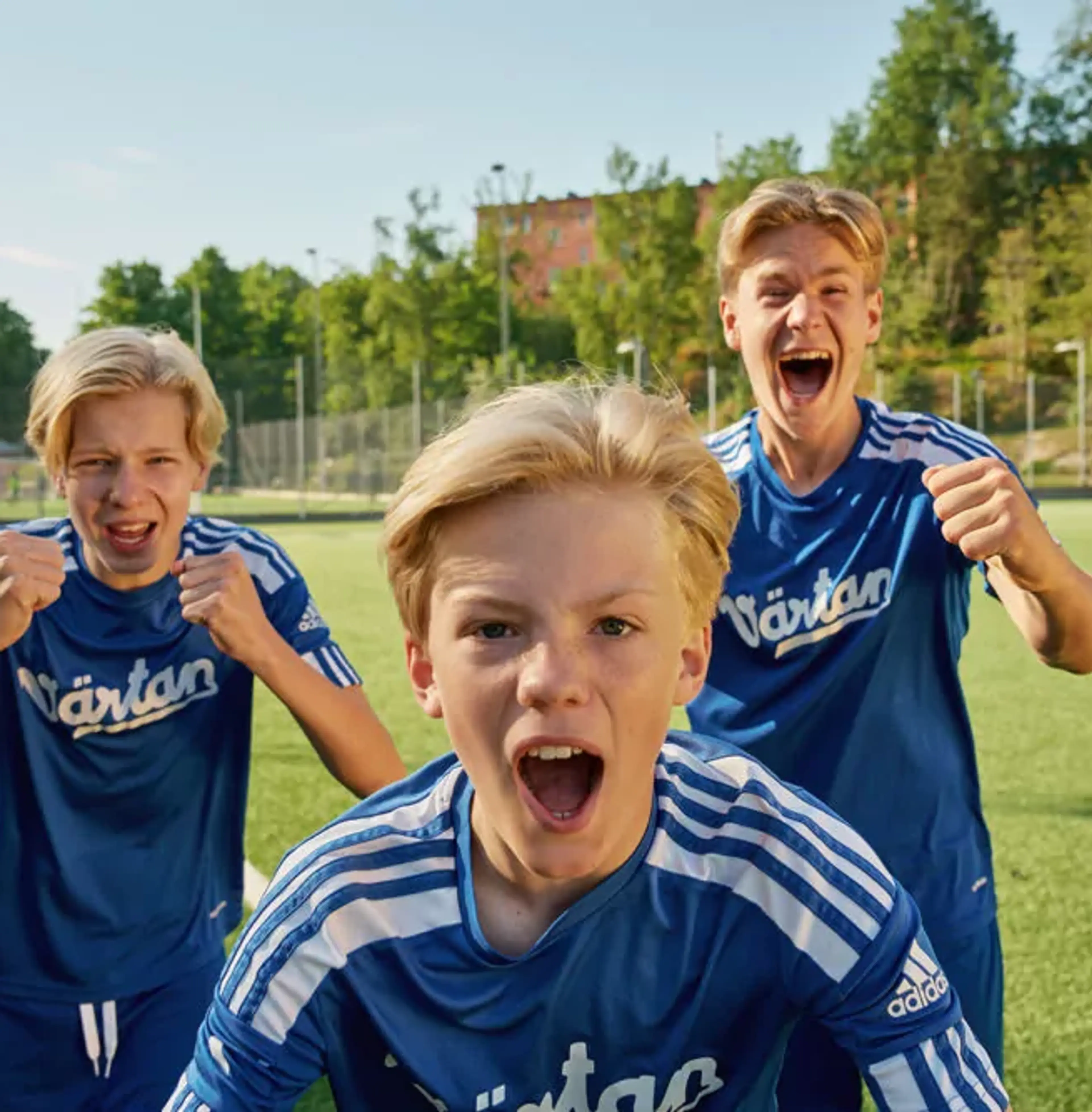 Tre pojkar i blå fotbollströjor står på en gräsplan och uttrycker glädje och pepp. Bakgrunden visar en solig dag med träd och byggnader.