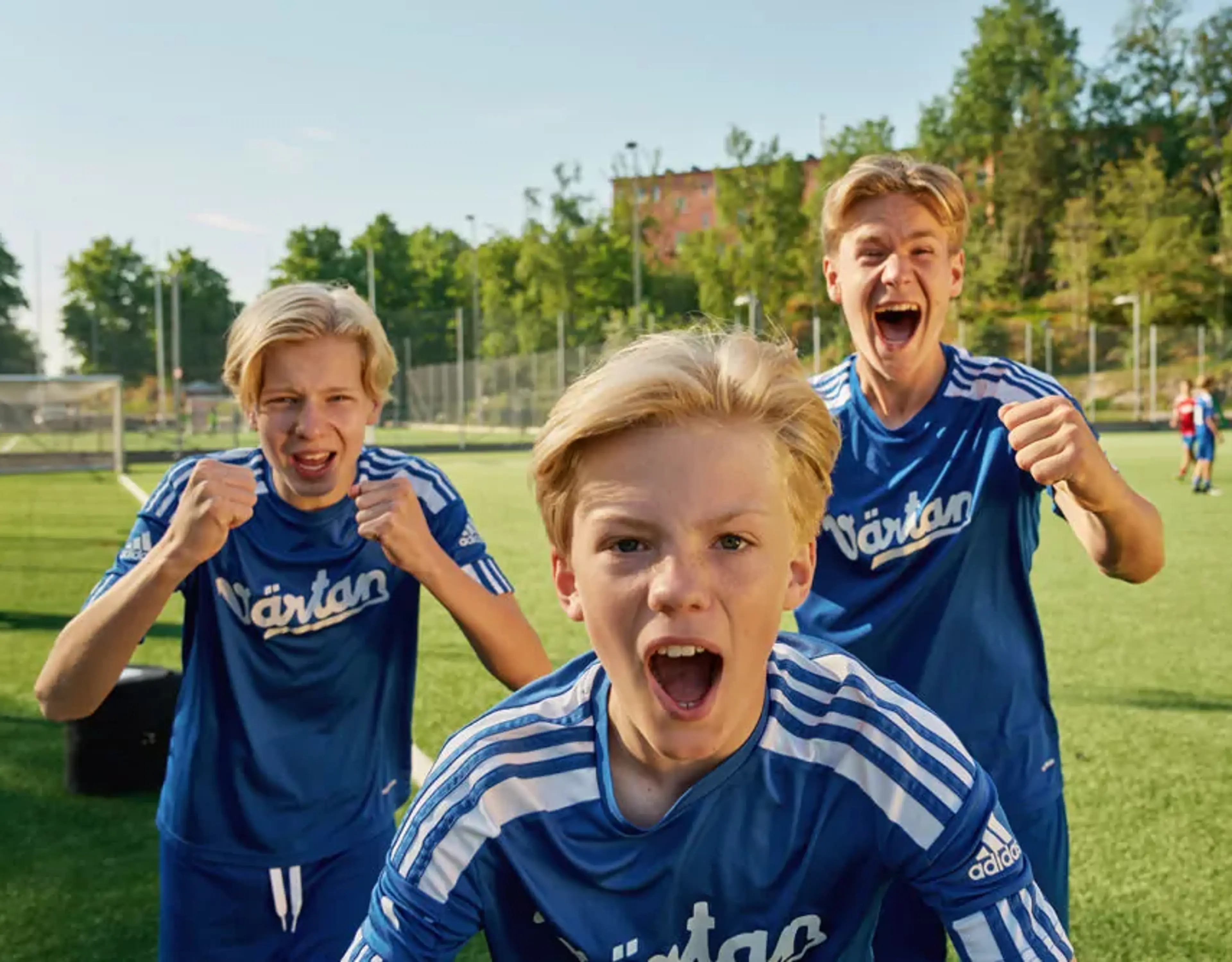 Tre pojkar i blå fotbollströjor står på en gräsplan och uttrycker glädje och pepp. Bakgrunden visar en solig dag med träd och byggnader.