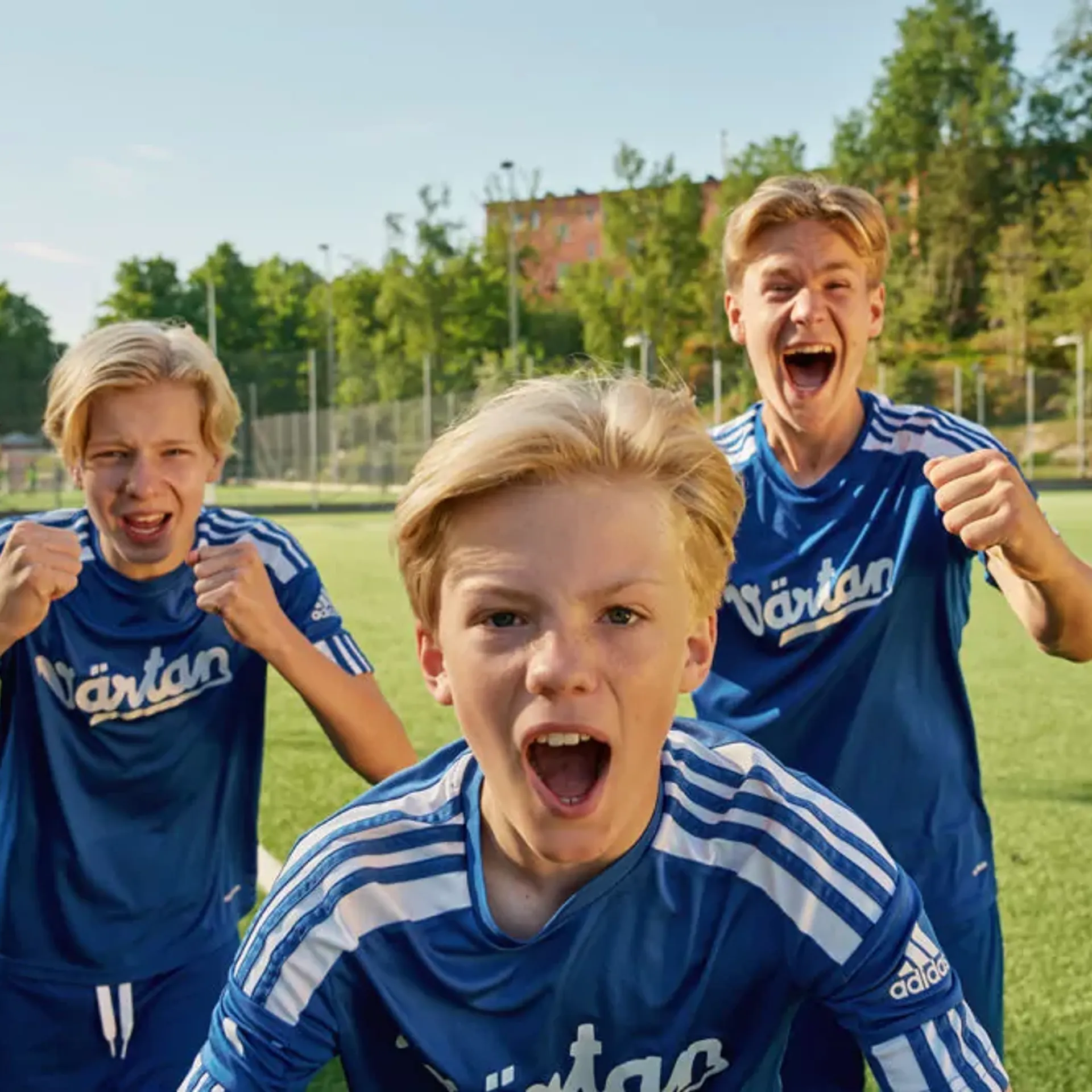 Tre pojkar i blå fotbollströjor står på en gräsplan och uttrycker glädje och pepp. Bakgrunden visar en solig dag med träd och byggnader.