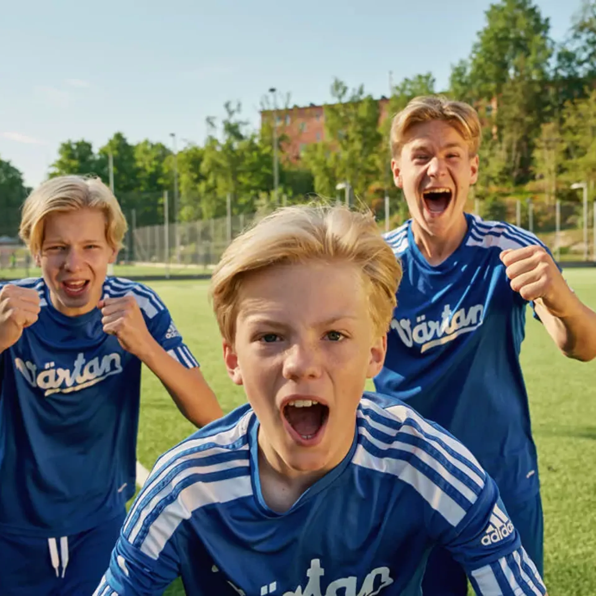 Tre pojkar i blå fotbollströjor står på en gräsplan och uttrycker glädje och pepp. Bakgrunden visar en solig dag med träd och byggnader.