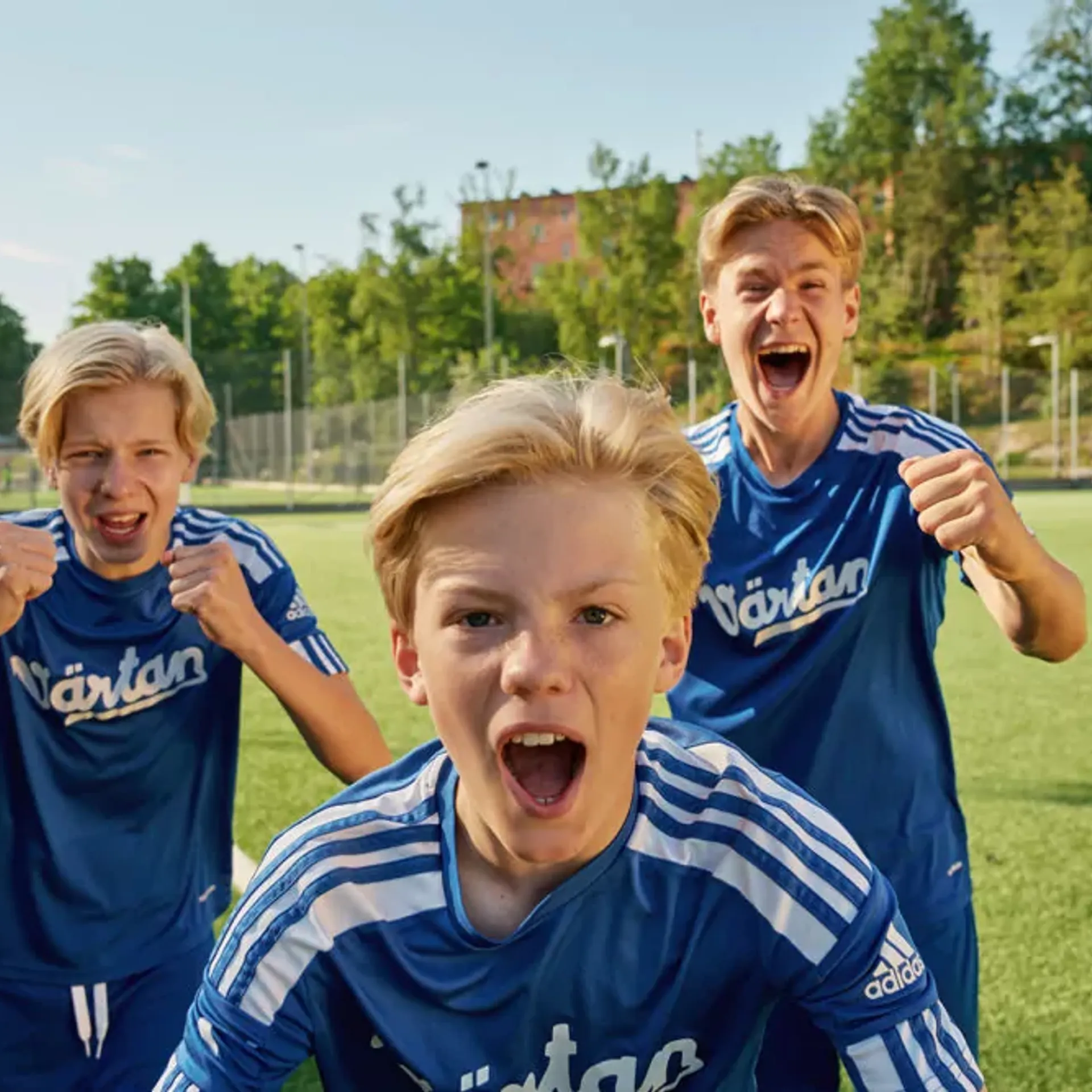 Tre pojkar i blå fotbollströjor står på en gräsplan och uttrycker glädje och pepp. Bakgrunden visar en solig dag med träd och byggnader.