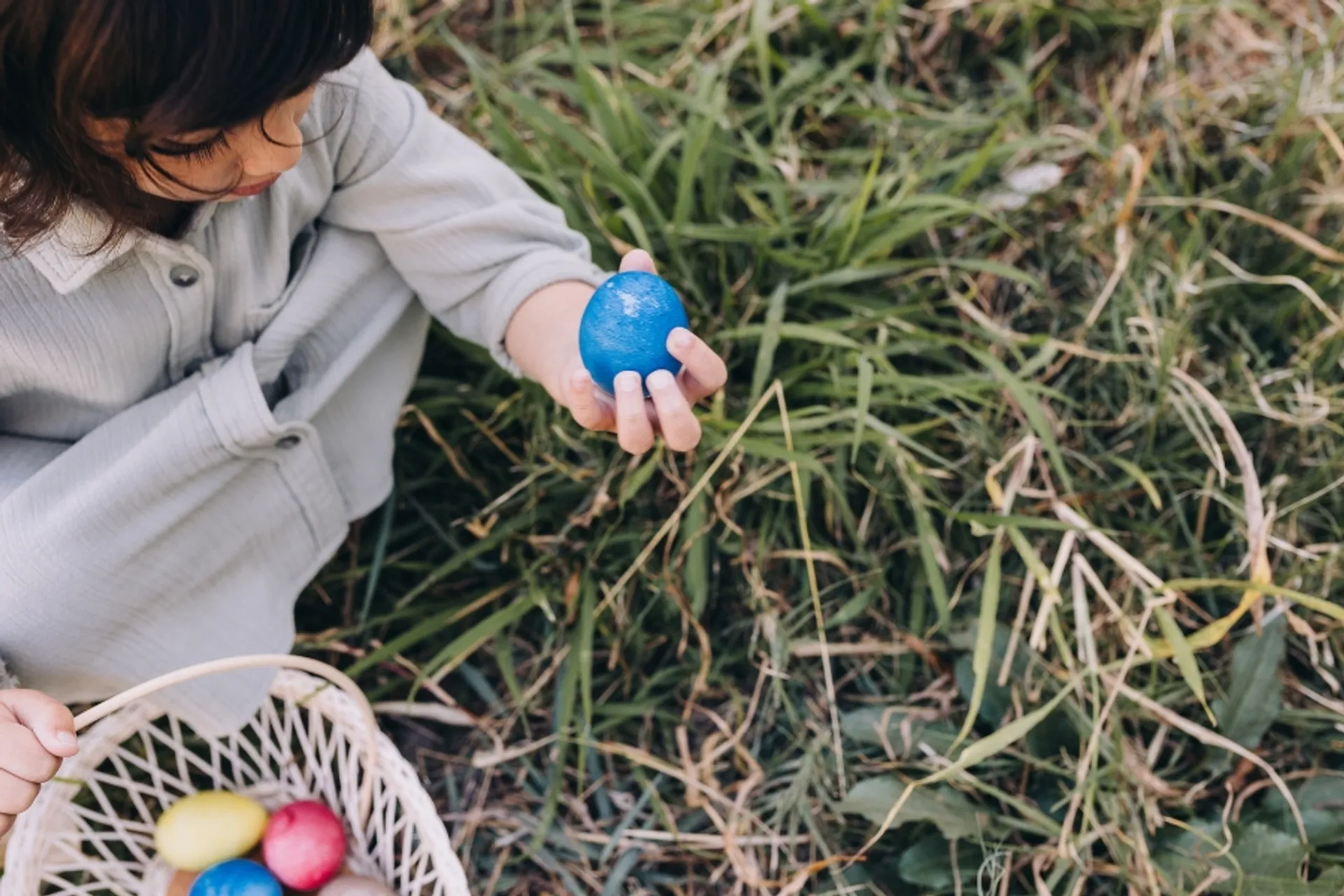 Ett barn sitter i gräset och håller i ett blått målat påskägg, medan en korg med fler färgglada ägg står intill.