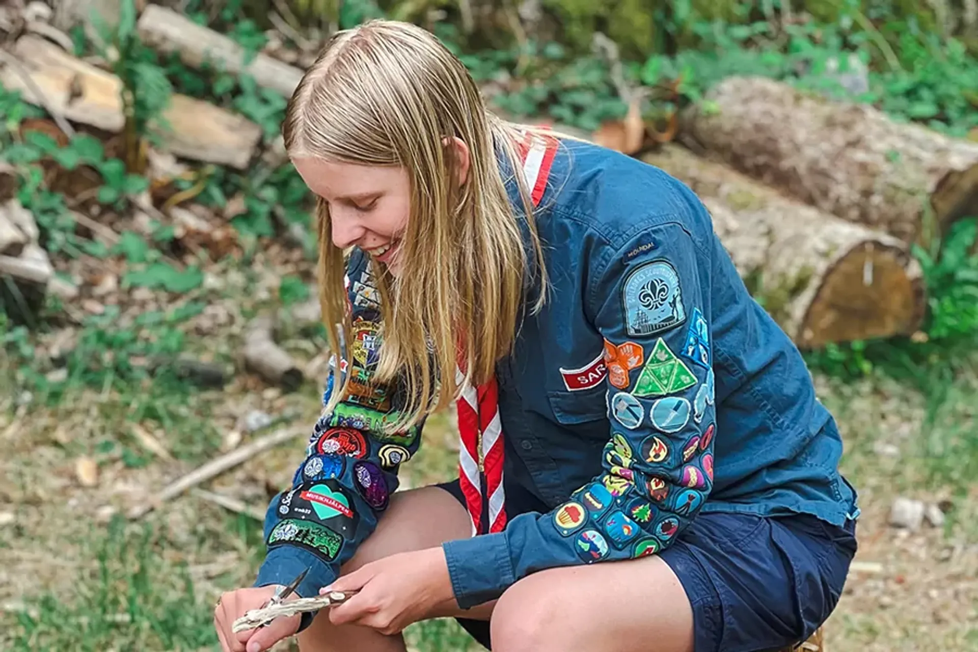 En ung scout med blå skjorta täckt av färgglada märken, som sitter ute i naturen och arbetar med en liten träbit. Bakgrunden visar en skogsmiljö med vedstockar och grönska.