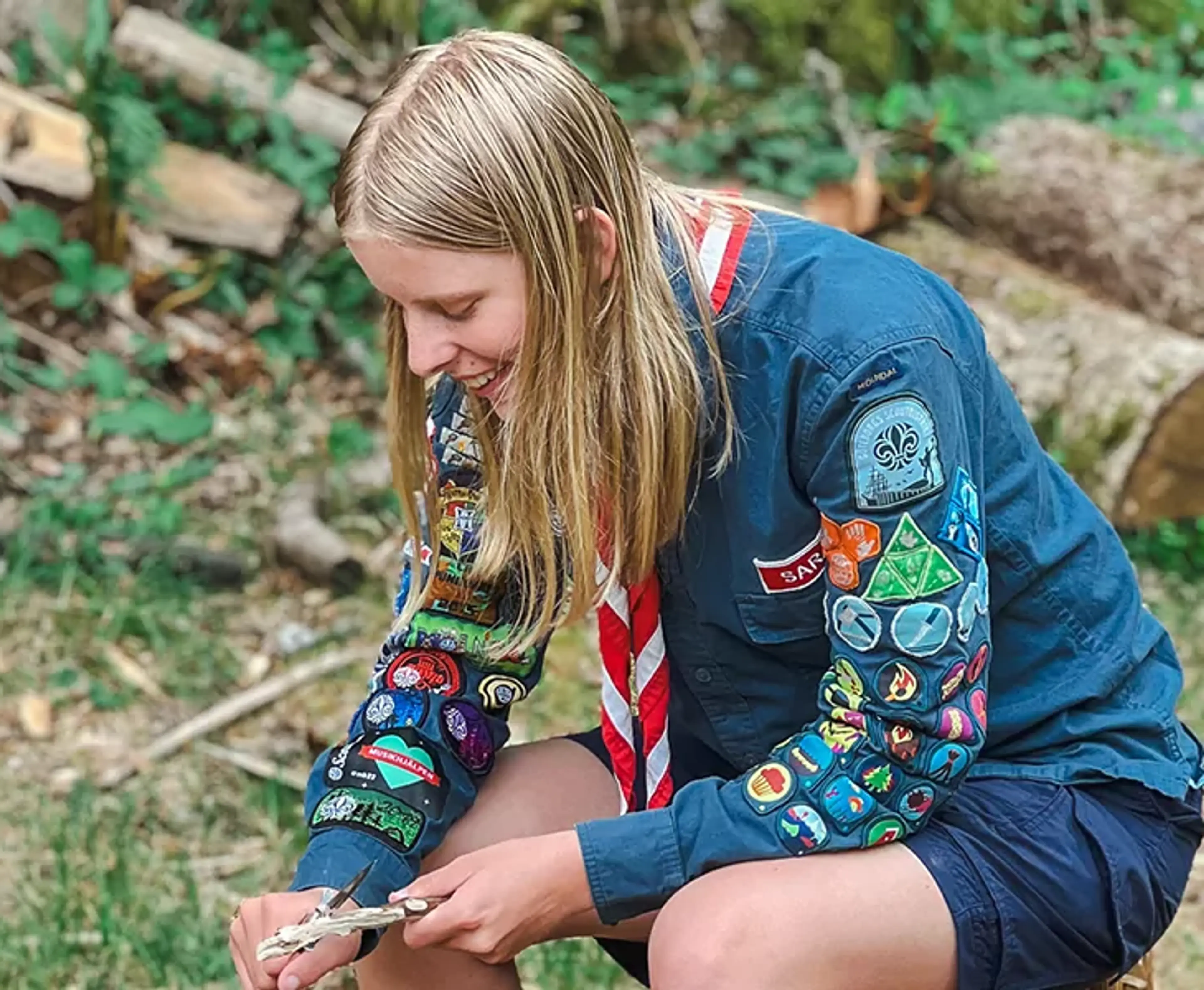En ung scout med blå skjorta täckt av färgglada märken, som sitter ute i naturen och arbetar med en liten träbit. Bakgrunden visar en skogsmiljö med vedstockar och grönska.
