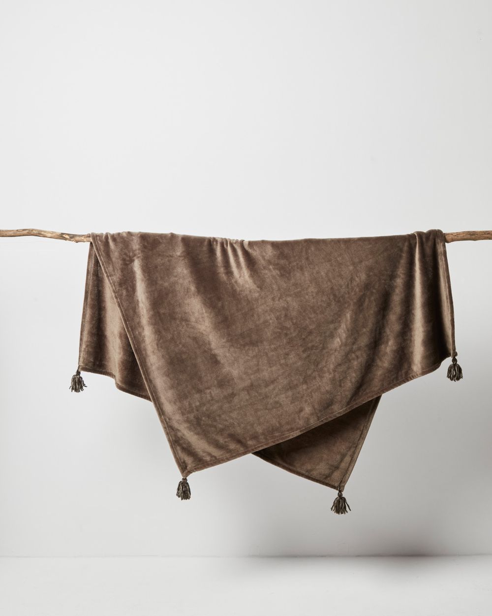 Mjuk fleecfilt i brun nyans med tofsar i hörnen, hängande över en tunn träpinne. Filten ser mysig och varm ut, perfekt för kyliga stunder.