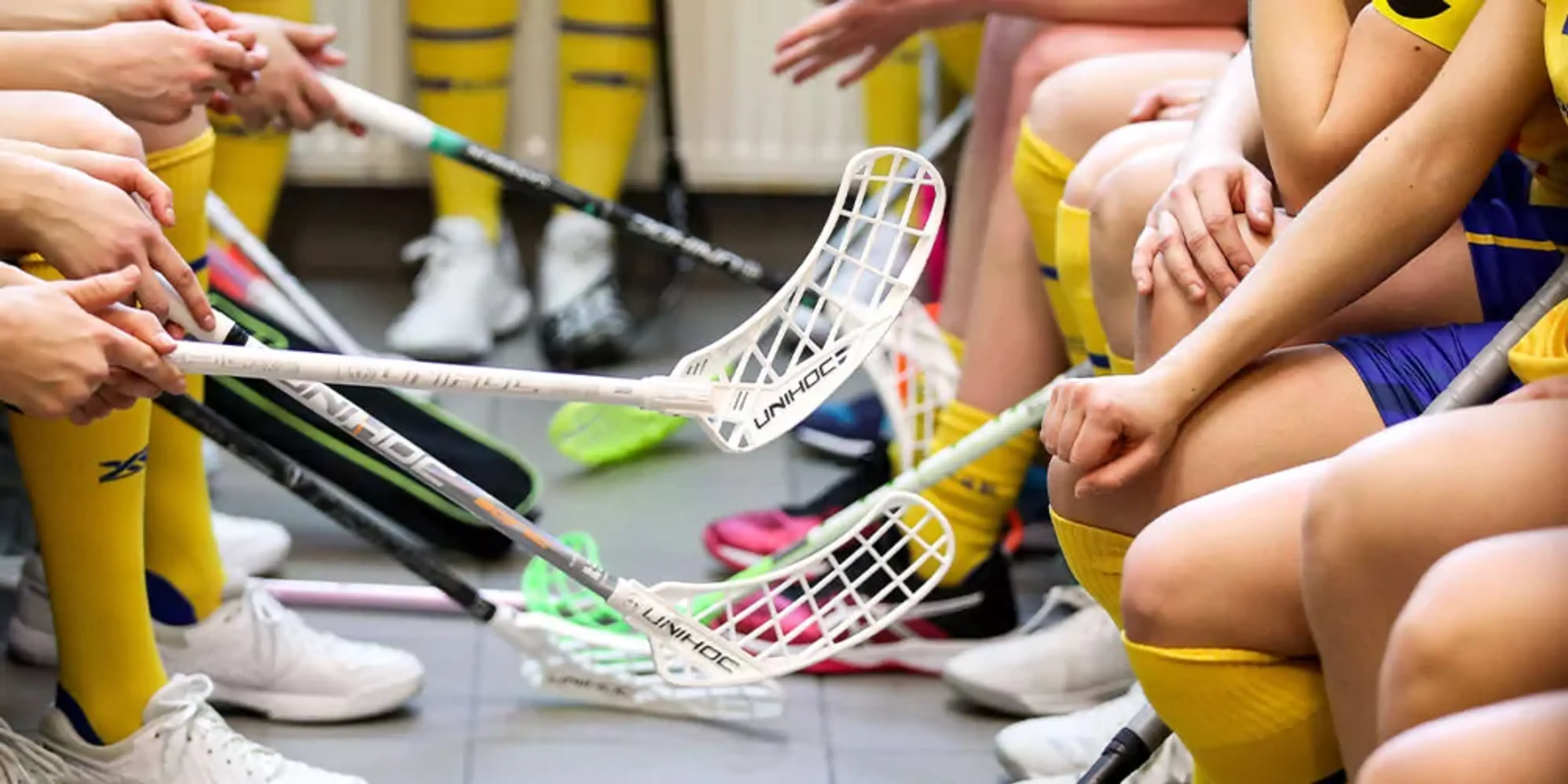 Handbollsklubbor och spelare i gula strumpor väntar tillsammans i en omklädningsrumsmiljö. Fokus ligger på klubborna som hålls nära golvet mellan två rader av spelare.