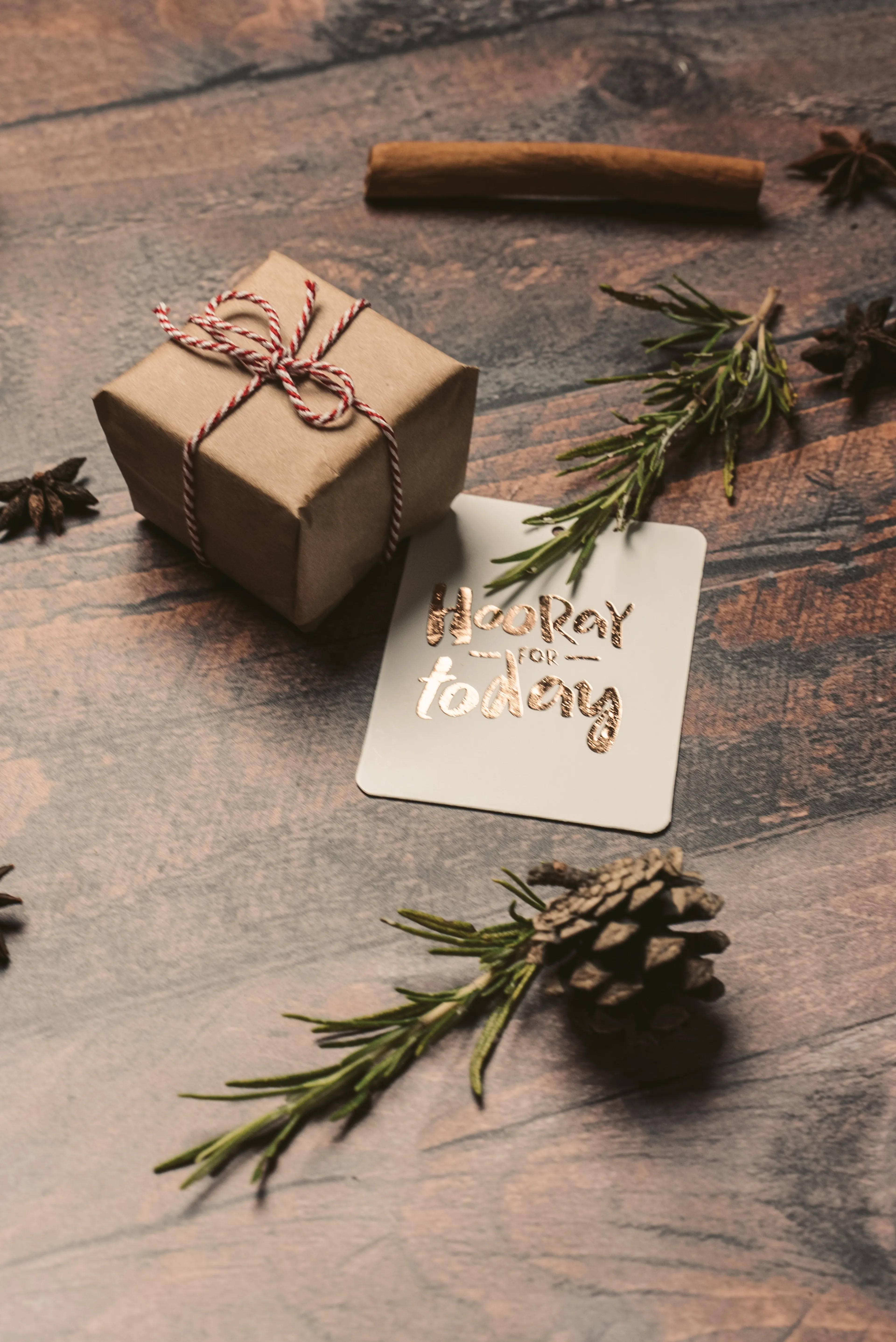 Ett litet paket inslaget i brunt papper och snöre ligger på ett träbord tillsammans med en grön kvist, en kanelstång, stjärnanis och en tänd etikett med texten "Hooray for today".