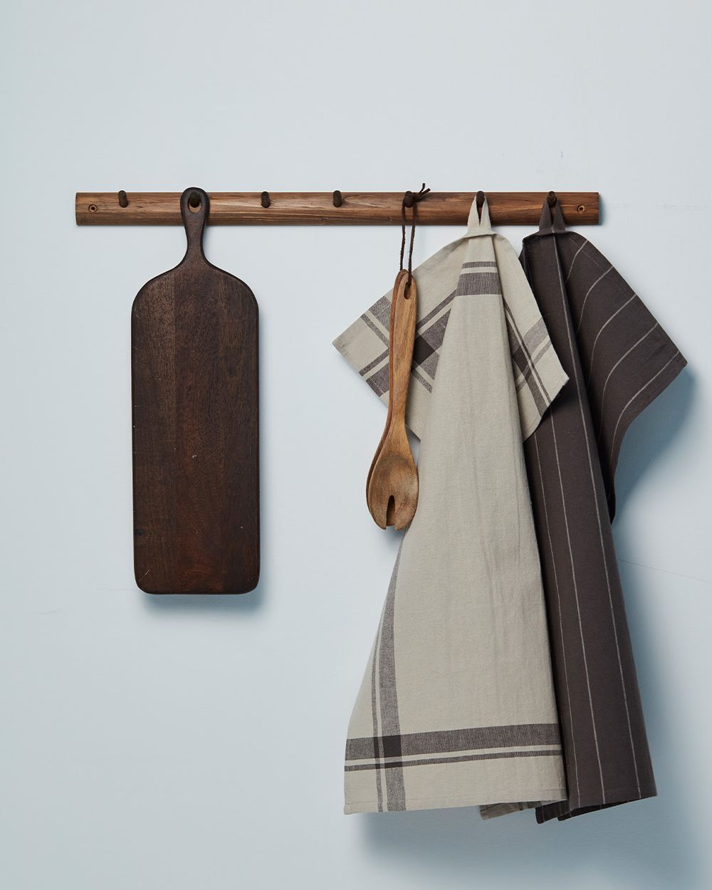 En träskärbräda, två randiga kökshanddukar i grått och beige samt en träslev hänger på en enkel, träfärgad kroklist mot en ljus vägg.