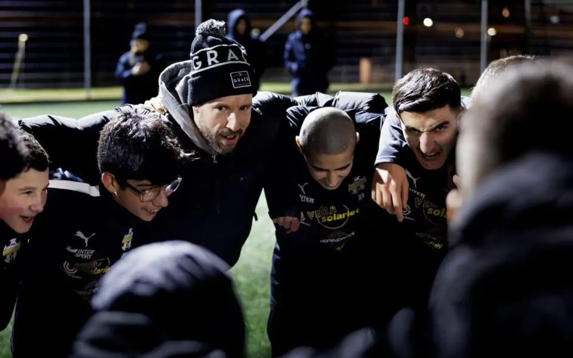 En grupp ungdomsspelare och en tränare i mössa står tätt ihop på en fotbollsplan under kvällstid, samlade i en peppande cirkel. De bär mörka idrottskläder med gula tryck.