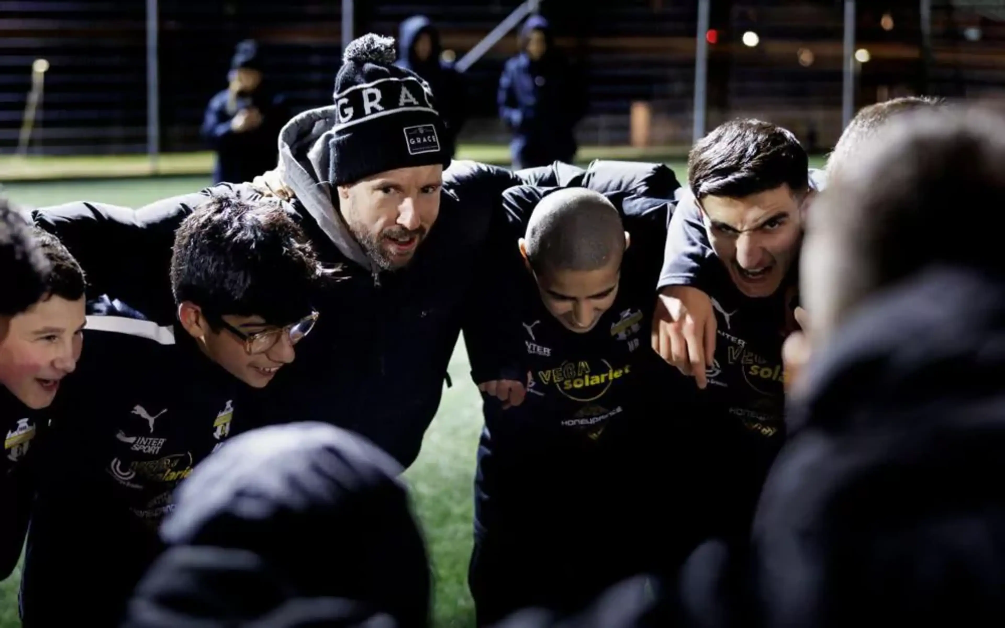 En grupp ungdomsspelare och en tränare i mössa står tätt ihop på en fotbollsplan under kvällstid, samlade i en peppande cirkel. De bär mörka idrottskläder med gula tryck.