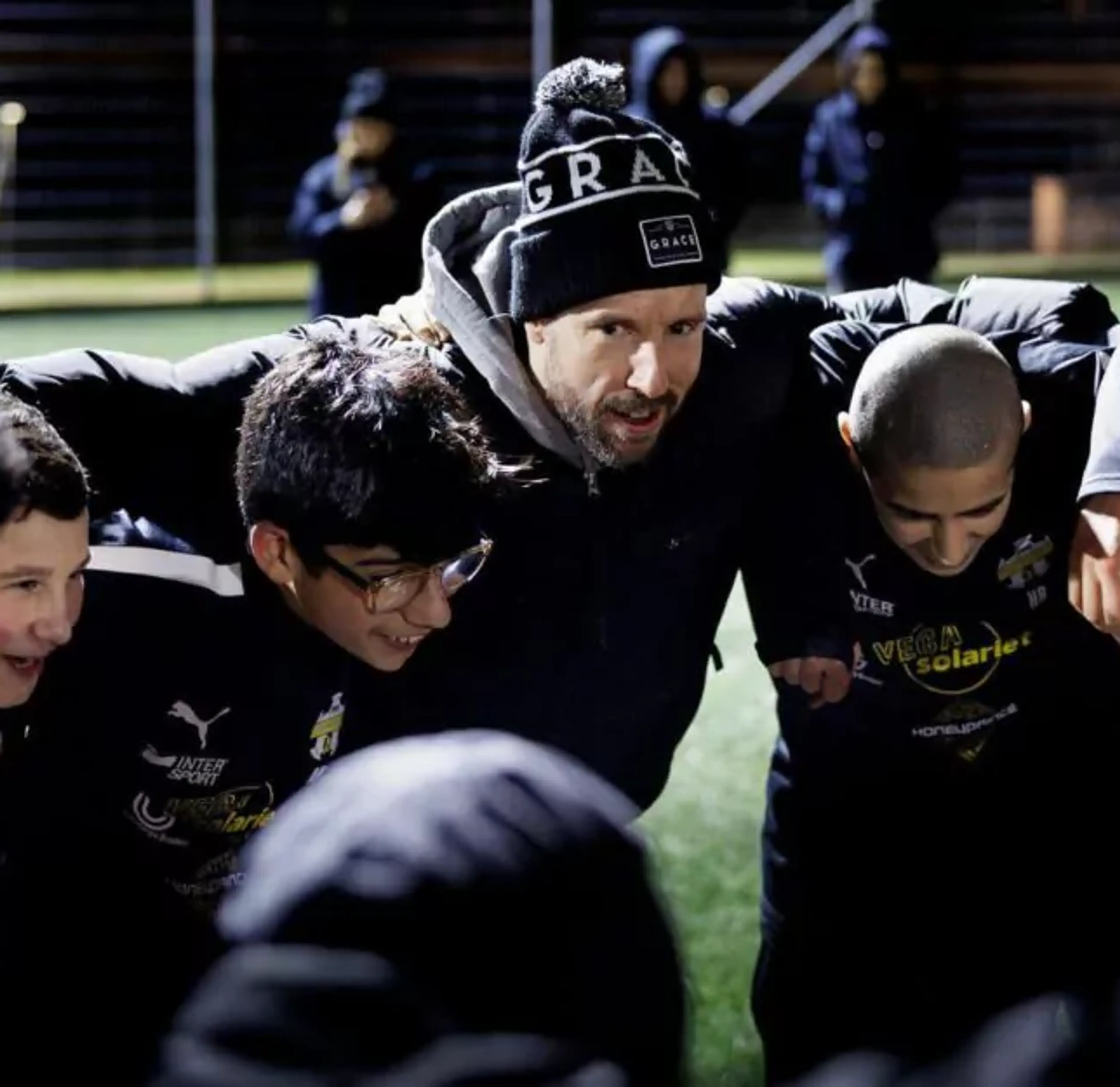 En grupp ungdomsspelare och en tränare i mössa står tätt ihop på en fotbollsplan under kvällstid, samlade i en peppande cirkel. De bär mörka idrottskläder med gula tryck.