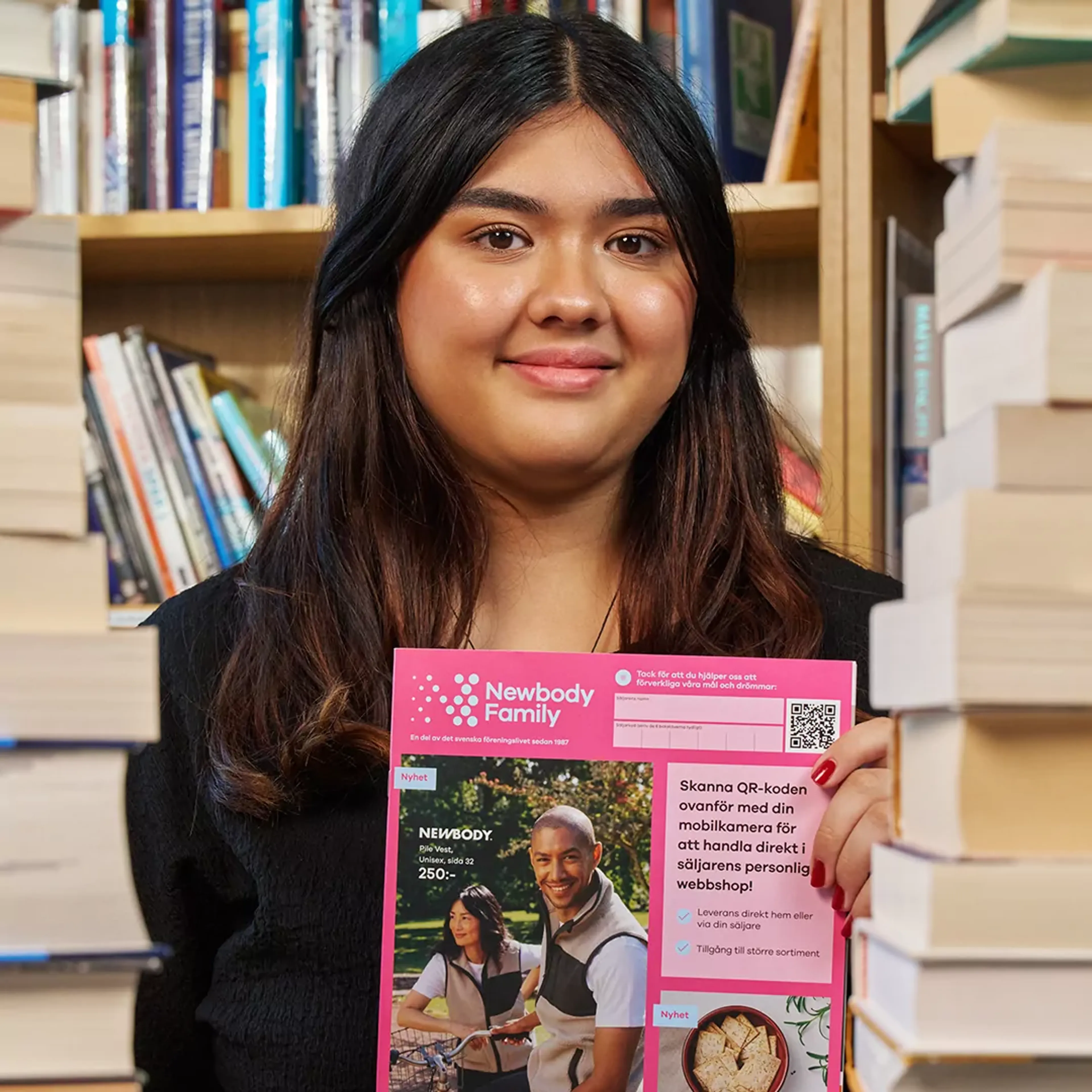 En ung tjej sitter mellan två högar med böcker och håller i en katalog med en rosa framsida från Newbody Family. I bakgrunden syns bokhyllor fyllda med böcker.